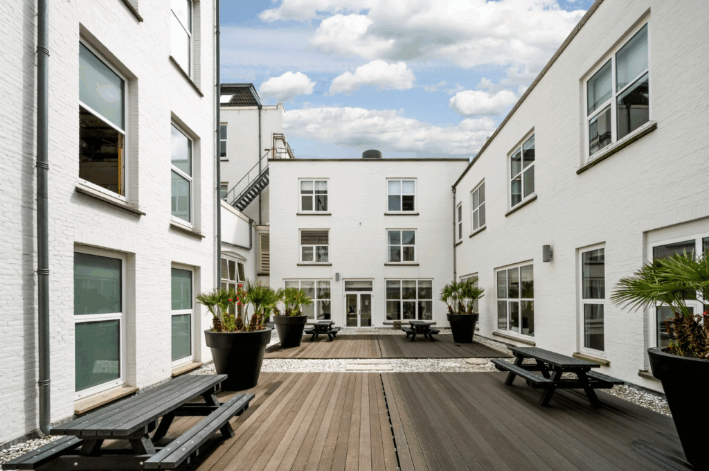 Moderne binnenplaats van de Paleisstraat met witte gebouwen, houten terrasplanken, zwarte picknicktafels en grote palmplanten in potten.