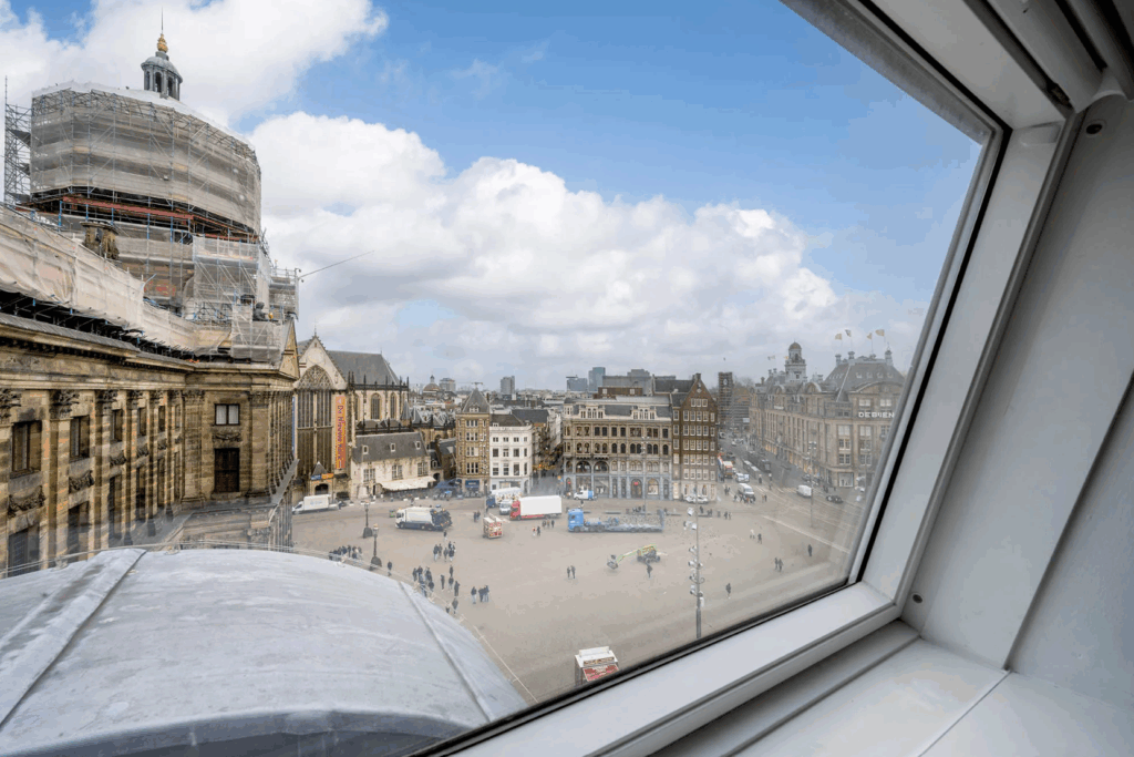 Uitzicht op de Dam in Amsterdam vanuit een raam, met het Koninklijk Paleis in renovatie en de omliggende historische gebouwen.