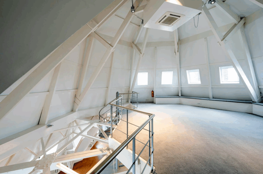 Ruime zolderkamer met schuine witte balken, kleine ramen en een metalen trap.