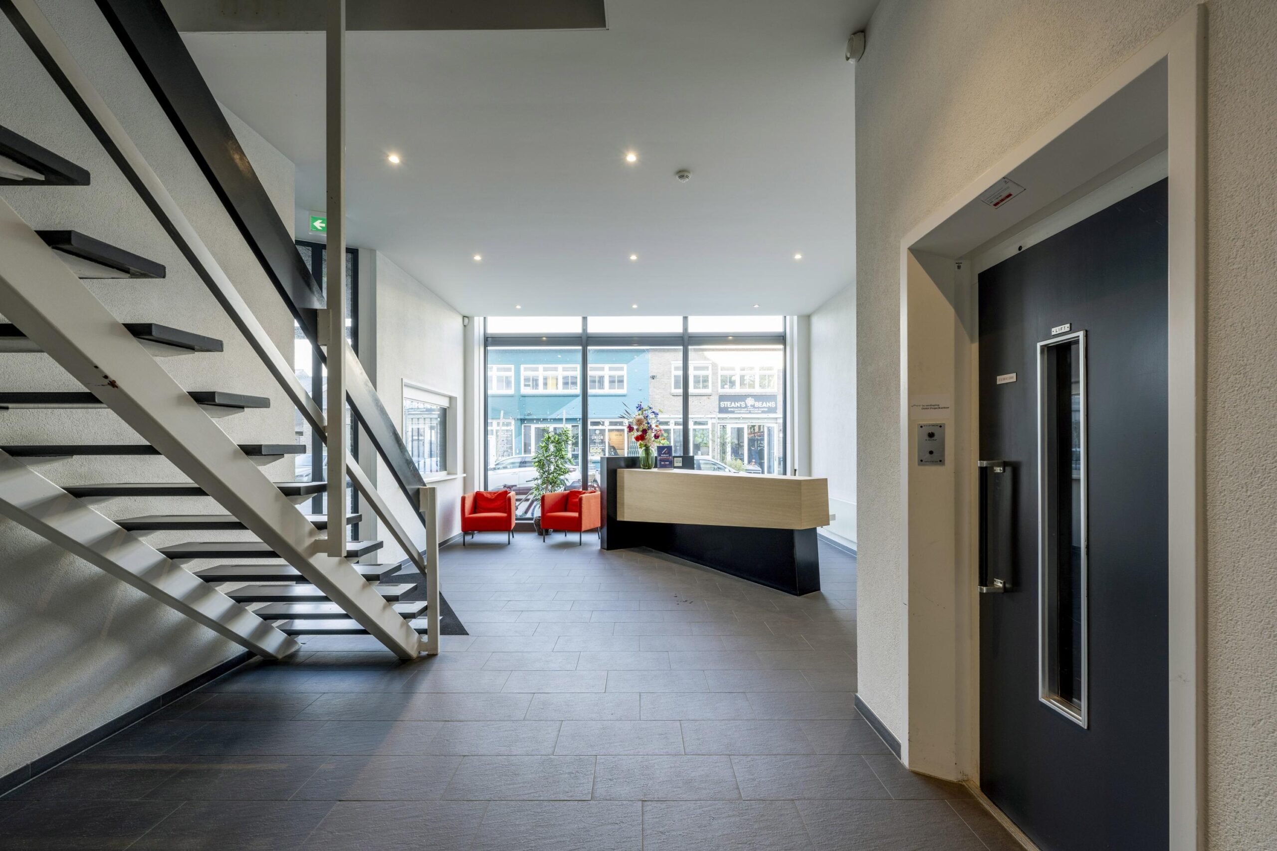 Moderne entreehal met receptiebalie, trap, lift en twee rode stoelen bij het raam aan de Helicopterstraat.