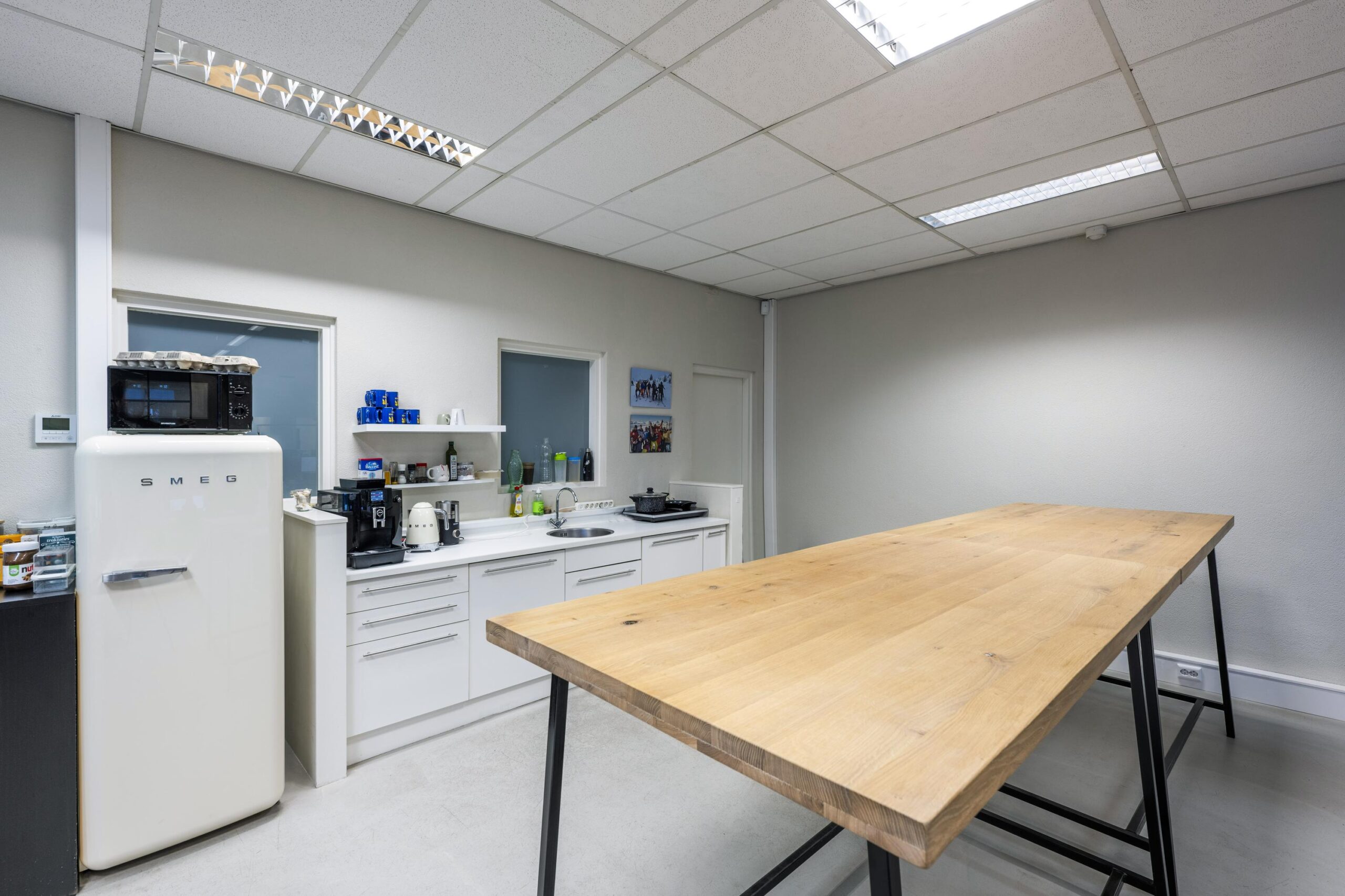 Moderne bedrijfskeuken aan de Helicopterstraat met witte kasten, houten tafel en Smeg-koelkast.