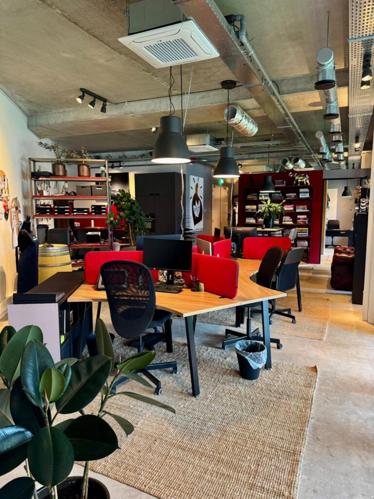 Modern open-plan office with shared wooden desks, red privacy screens, and industrial-style ceiling.