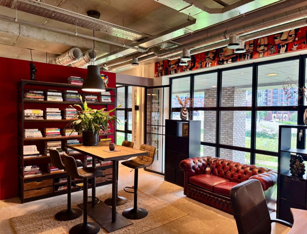 Modern office lounge with a red leather Chesterfield sofa, tall bookshelves, and large industrial-style windows.