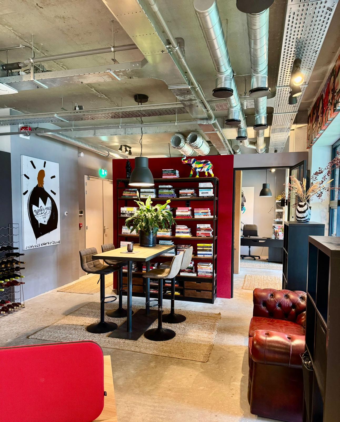 Modern ingerichte kantoorruimte met een hoge tafel, boekenkast, leren fauteuil en industriële plafondbuizen.