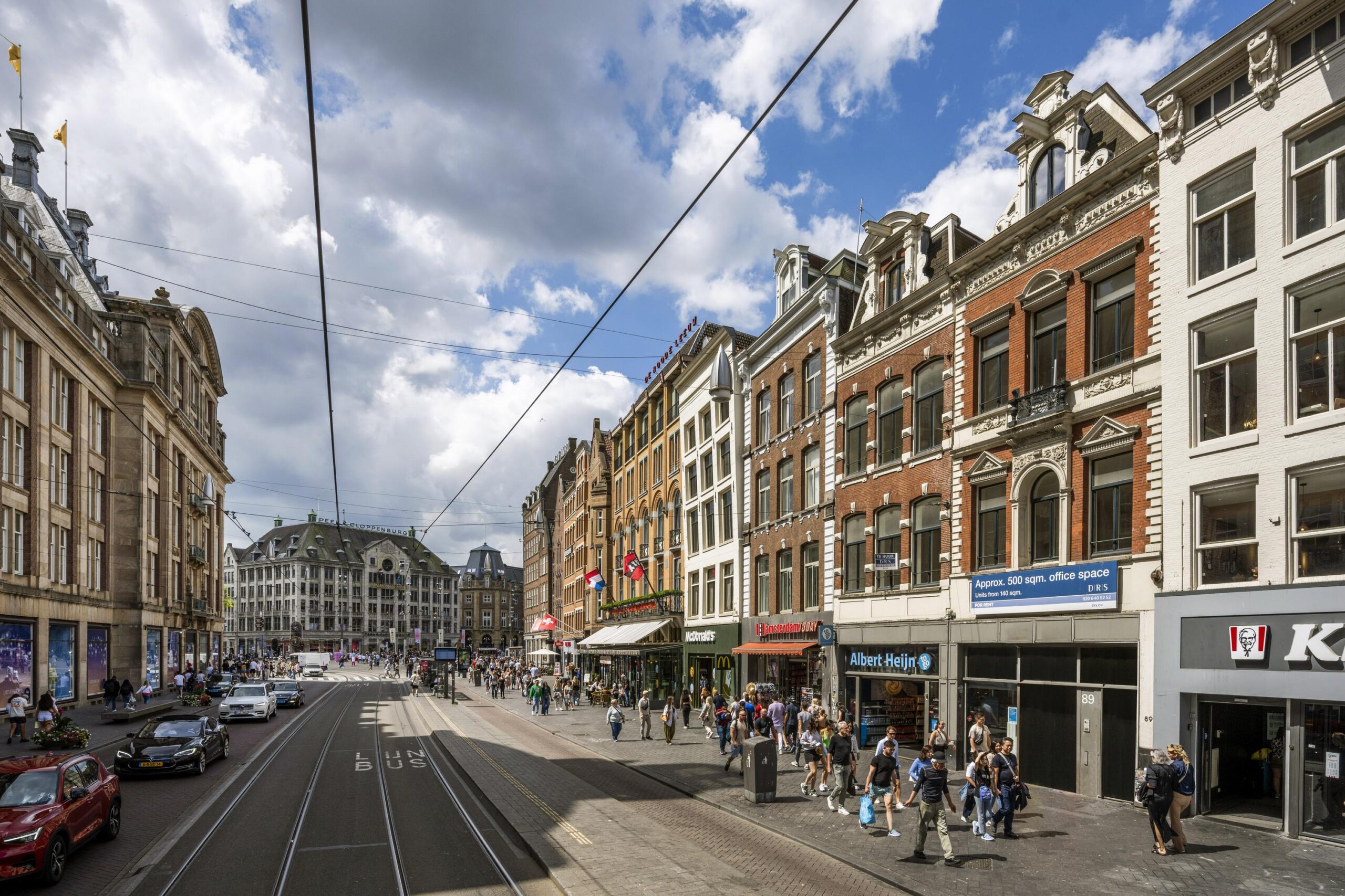 Drukke winkelstraat Damrak in Amsterdam met historische gebouwen, trambaan en winkelend publiek.