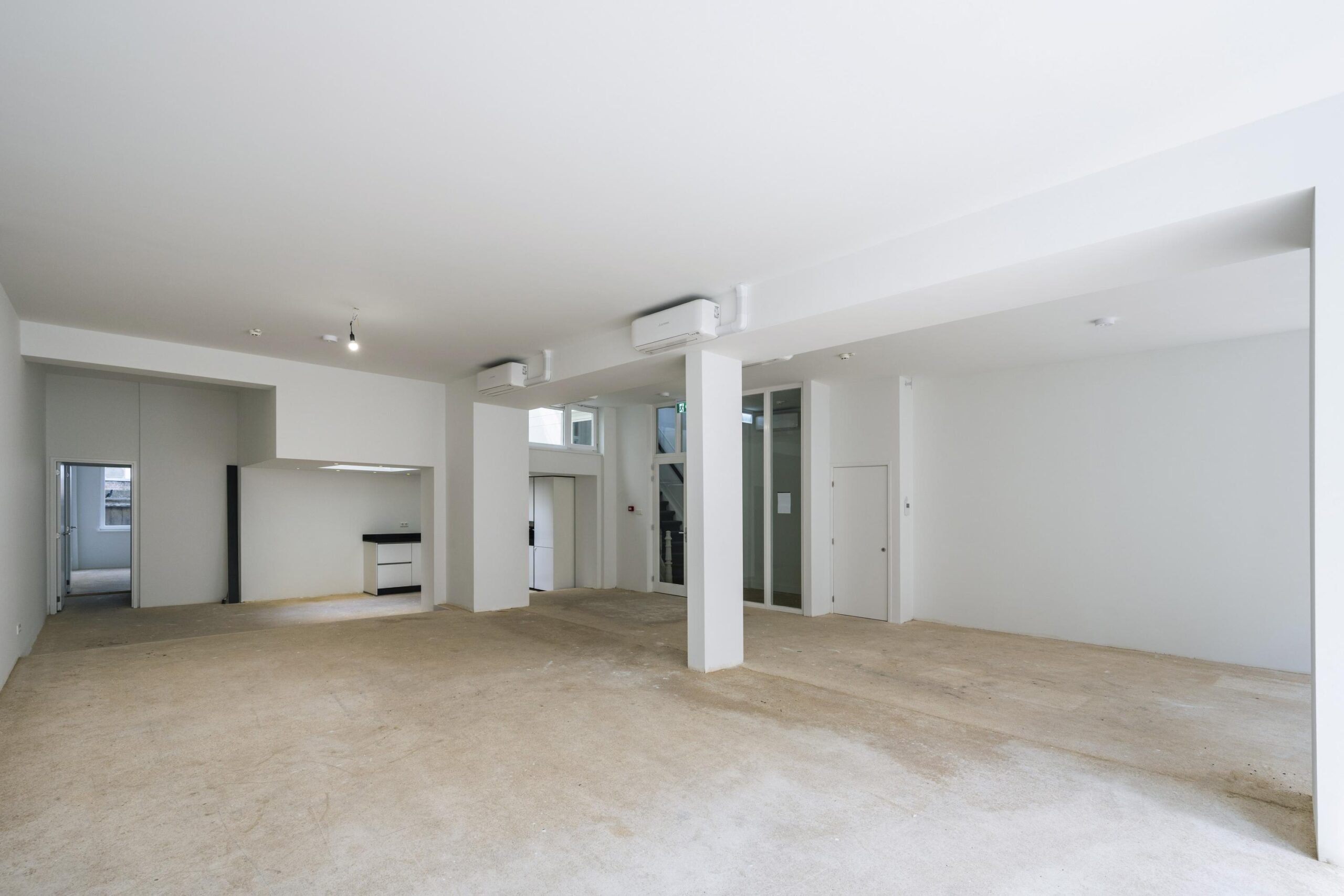 Leegstaand commercieel interieur aan het Damrak met witte muren, betonnen vloer en zicht op een kitchenette en trappenhuis.