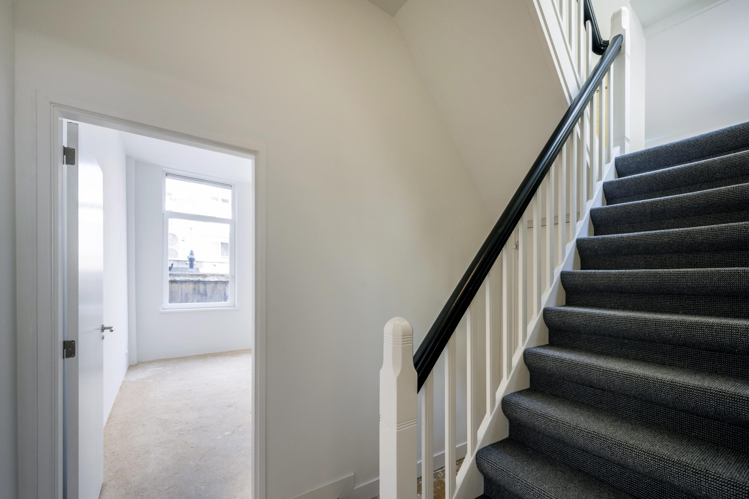 Trapopgang met donkergrijze vloerbedekking en een open deur naar een lichte kamer aan het Damrak.