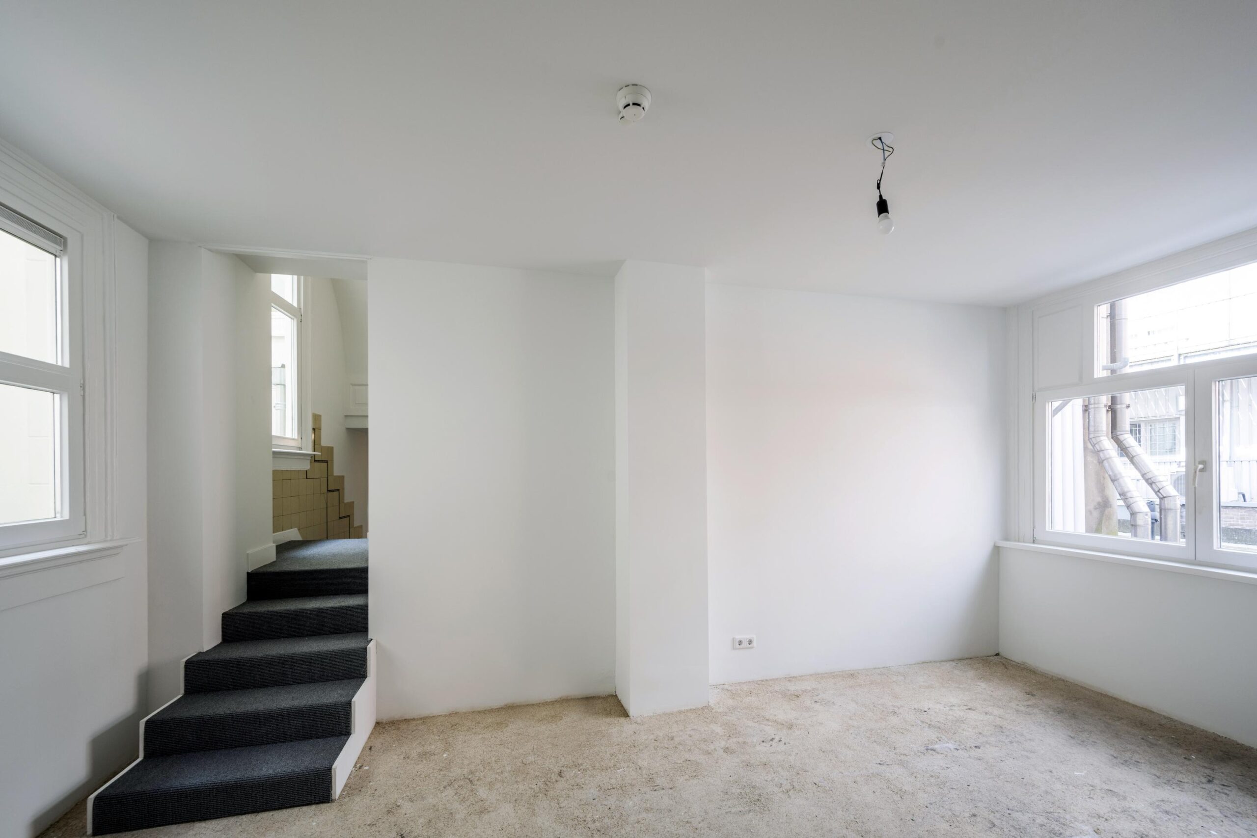 Lege, witgeschilderde kamer aan het Damrak met onafgewerkte vloer, trap en grote ramen.