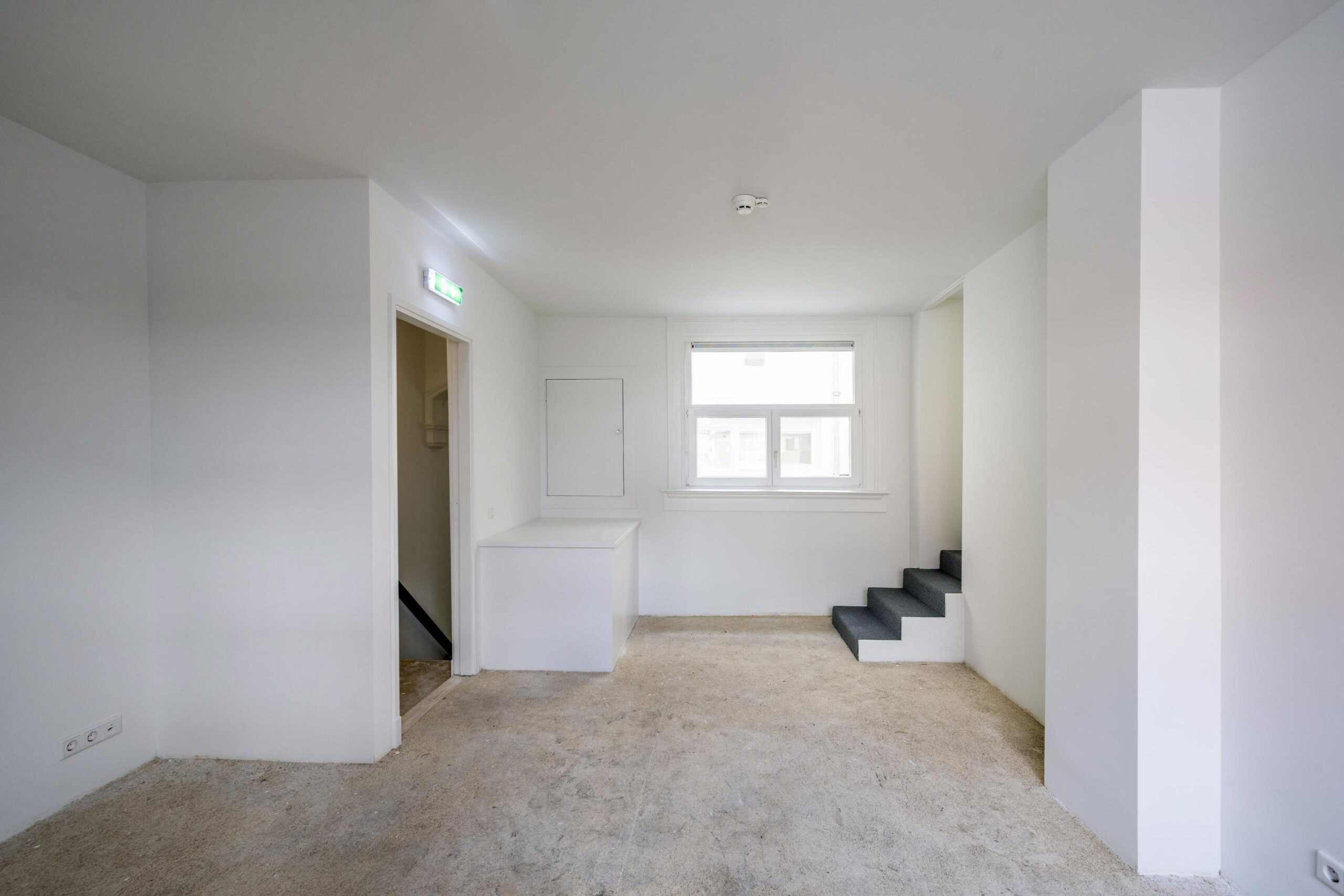 Leeg, witgeschilderd interieur met een trapje, raam en deuropening in een ruimte aan de Damrak.
