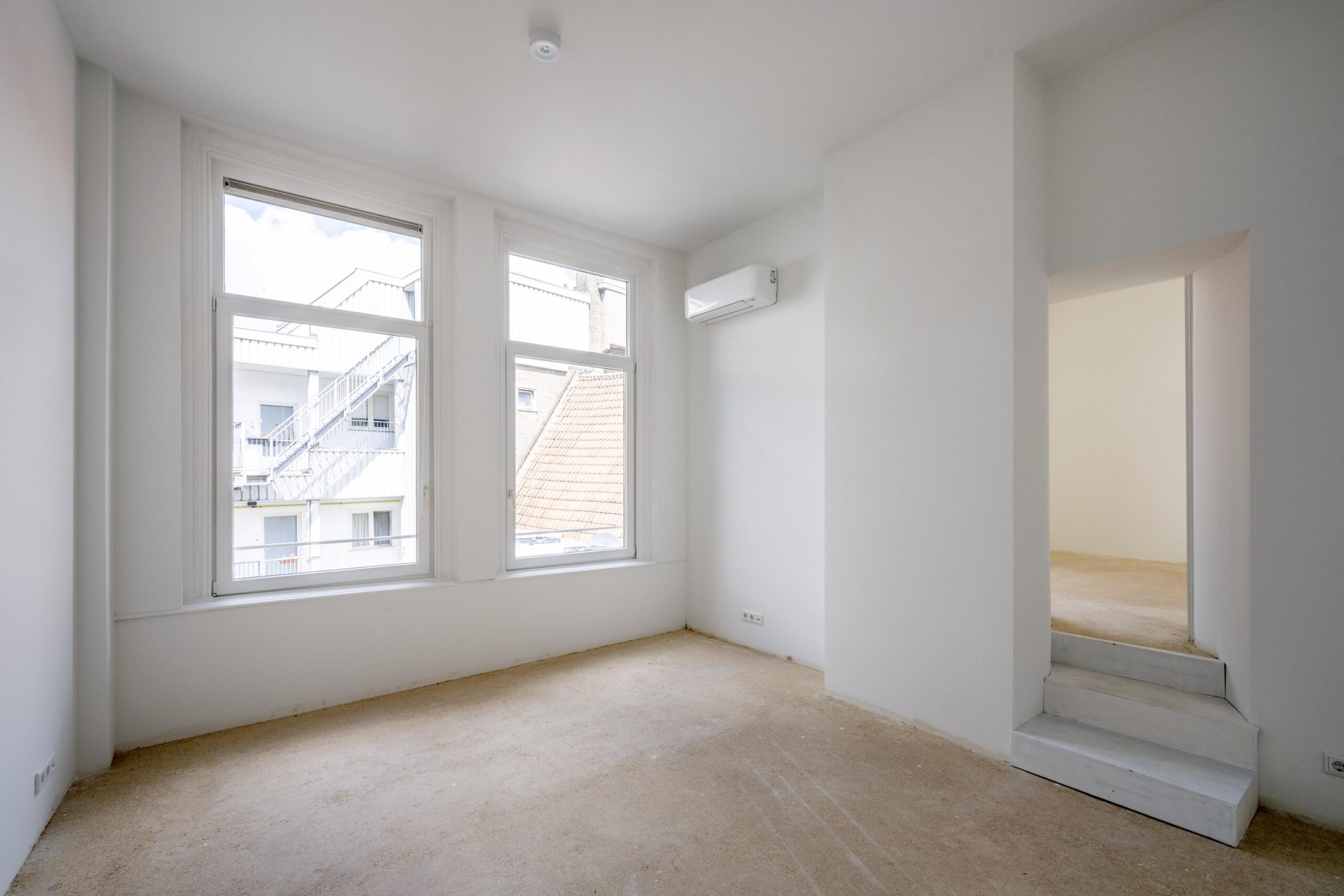 Licht lege kamer aan het Damrak met grote ramen, witte muren en zicht op aangrenzende gebouwen.