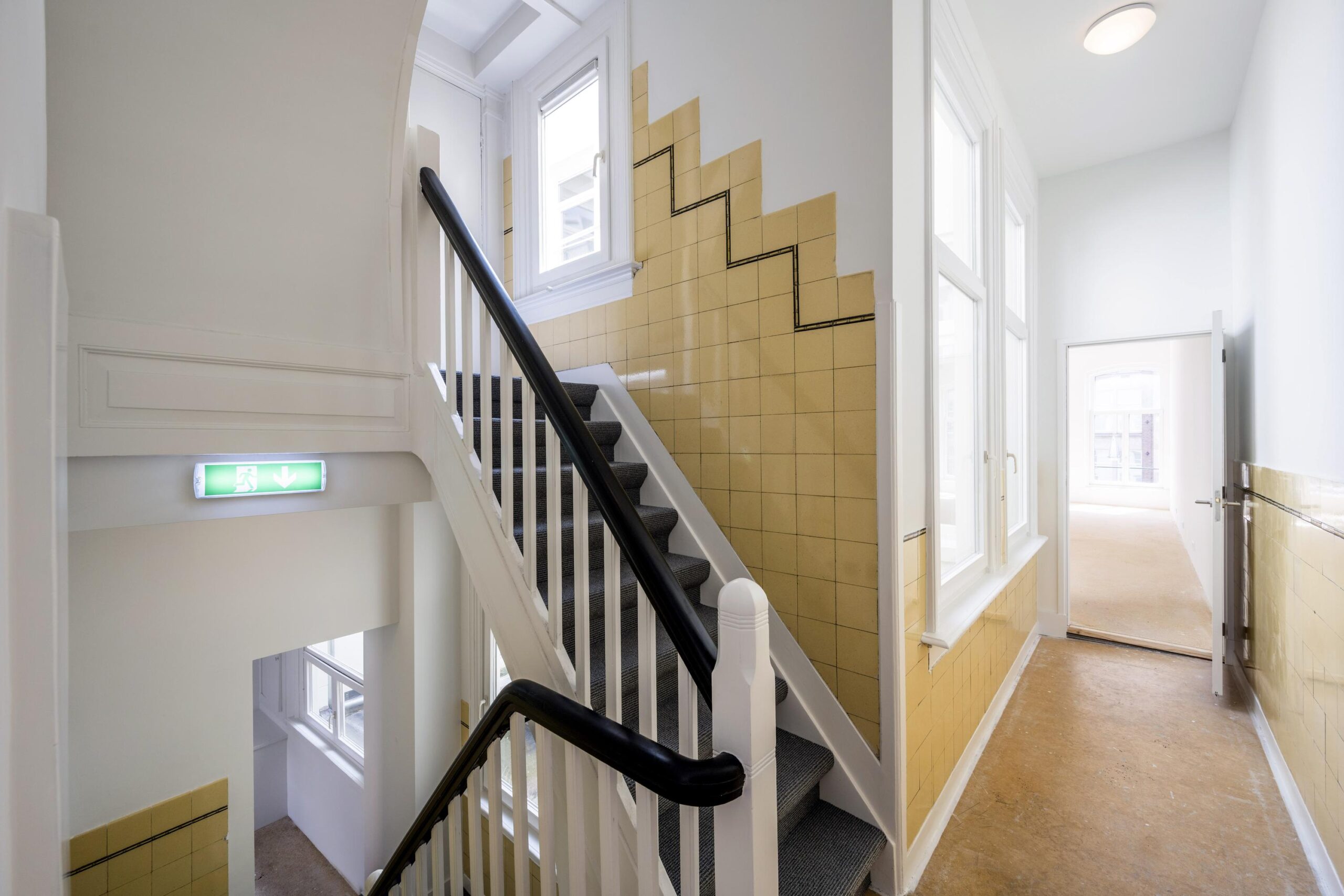 Trapportaal in een pand aan het Damrak met beige wandtegels, witte muren en natuurlijke lichtinval.