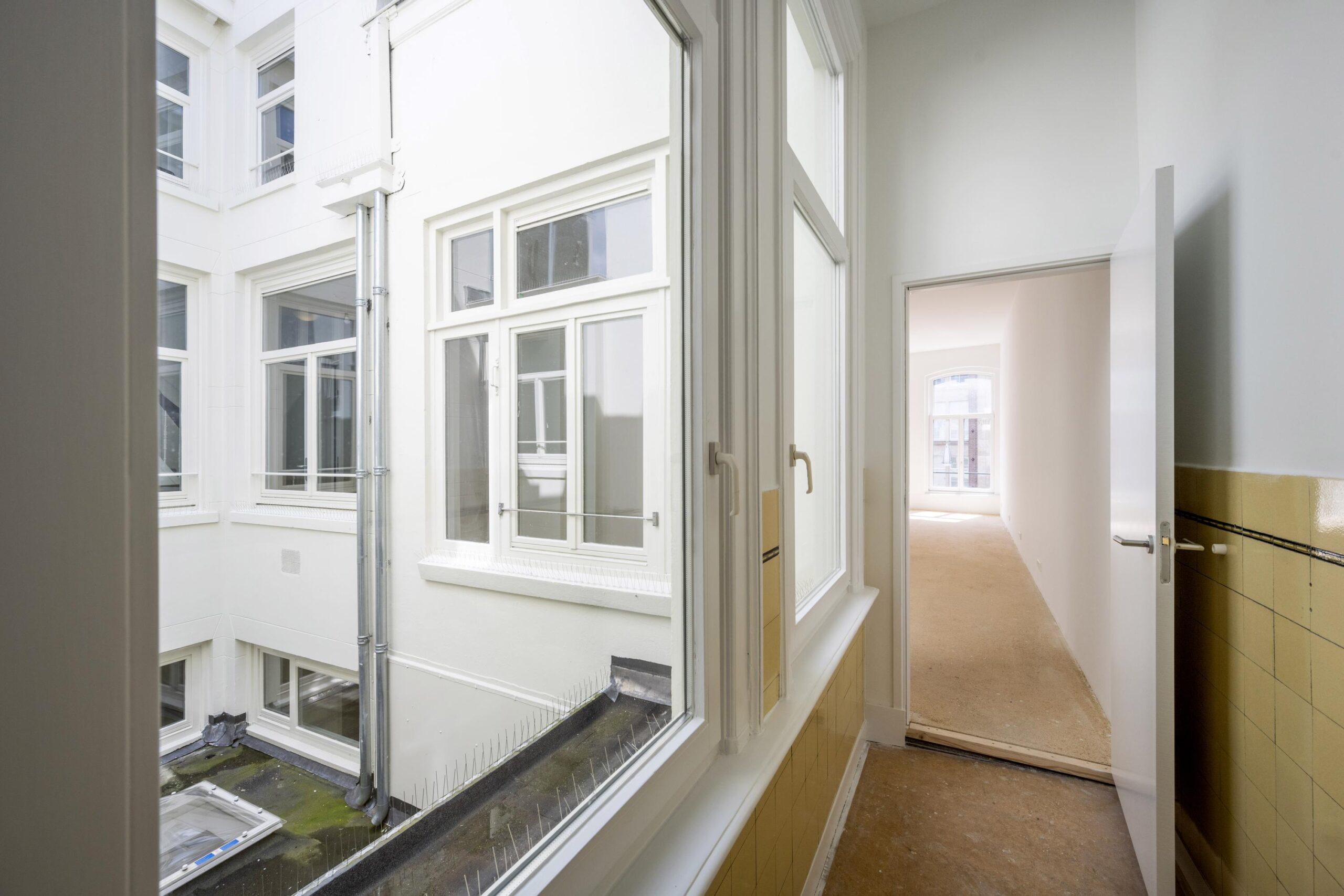 Uitzicht vanuit een gang of kamer op een binnenplaats met meerdere witte ramen en een open deur naar een lichte ruimte.