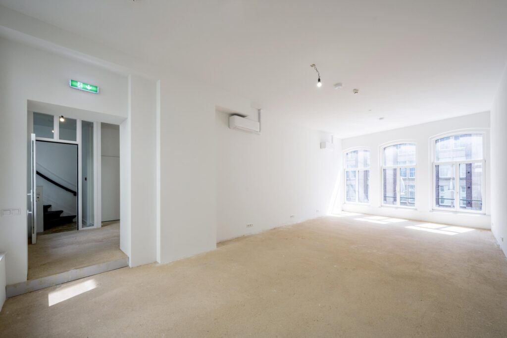 Lichte, lege ruimte op de Damrak met grote ramen en een onafgewerkte vloer.