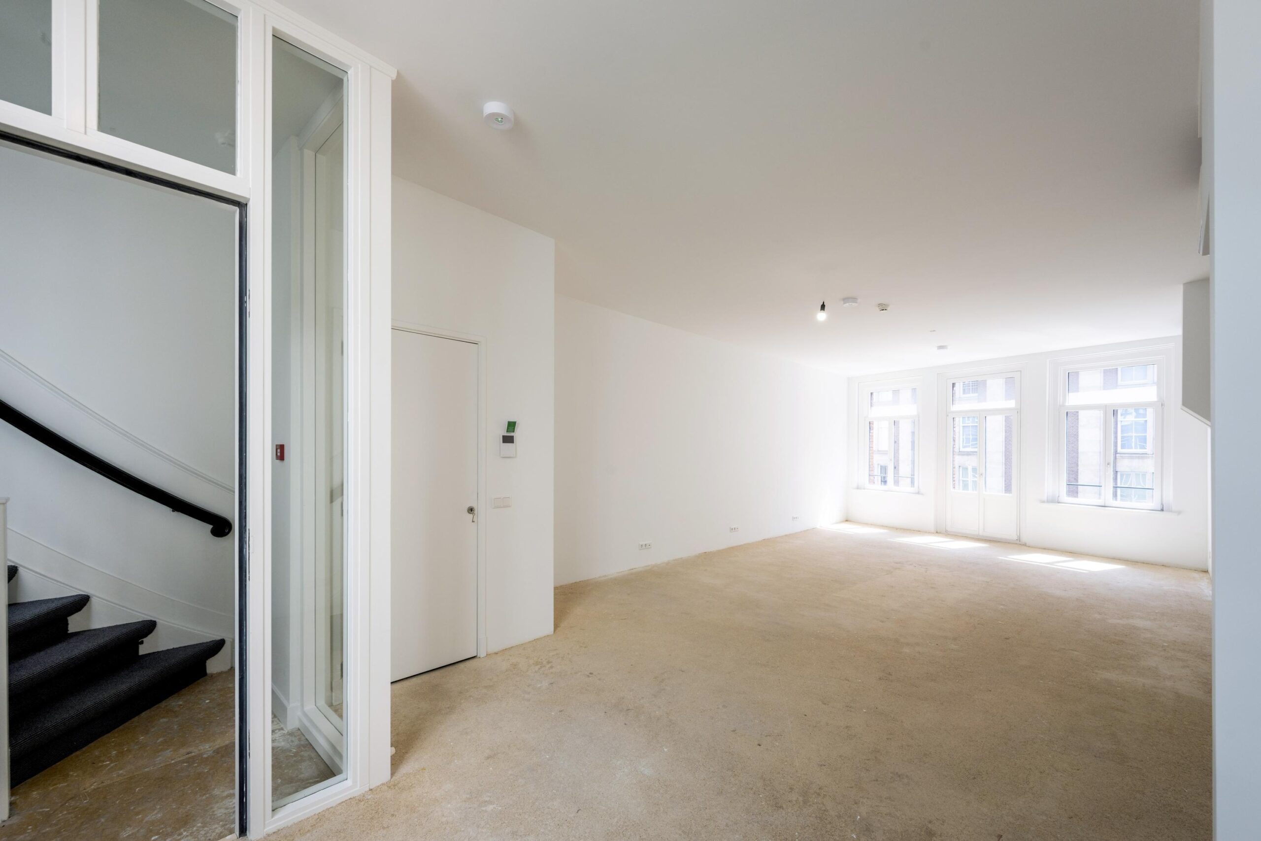 Lichte, lege woonruimte aan het Damrak met grote ramen en zicht op een trapopgang.