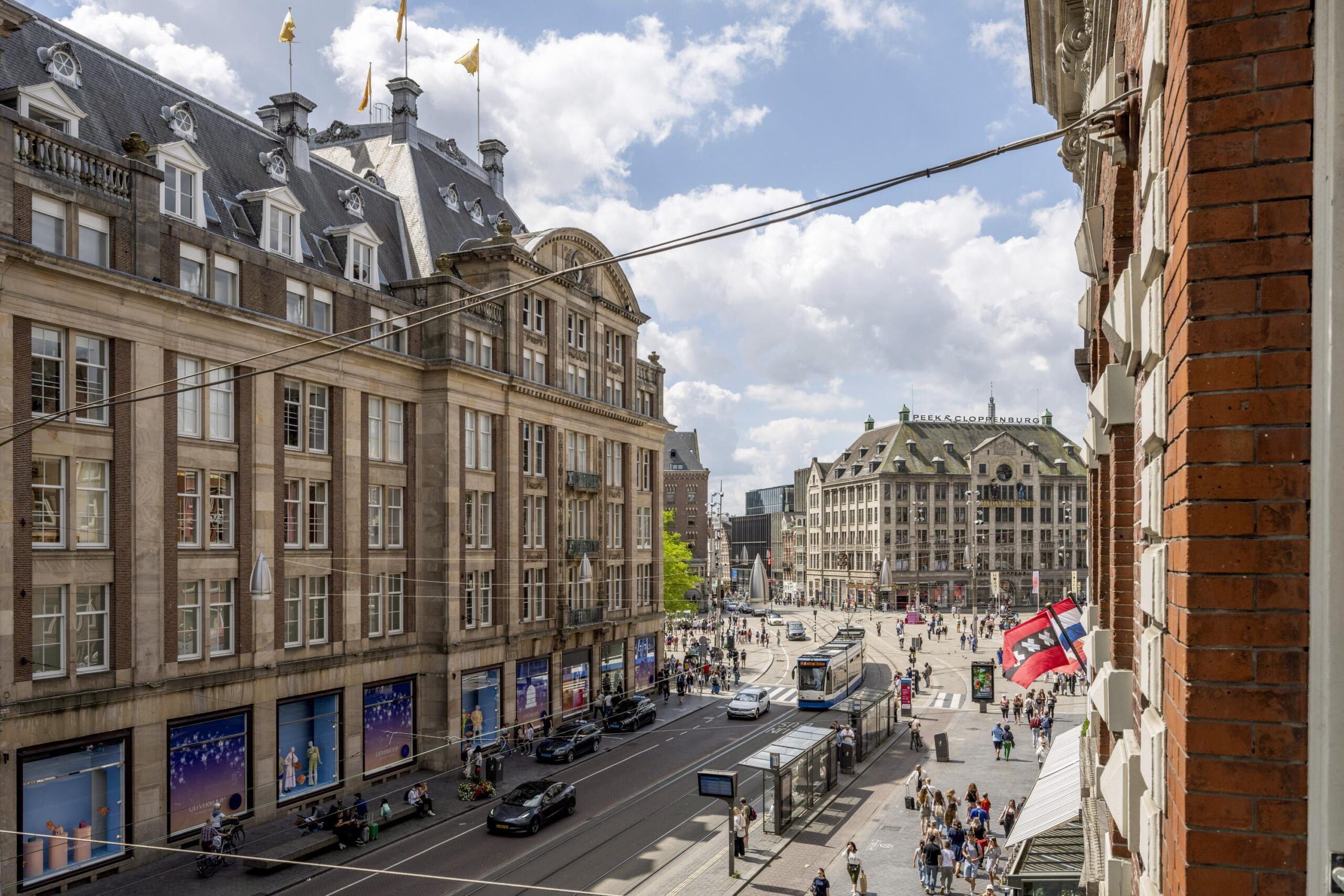 Drukke straatscene op het Damrak in Amsterdam met historische gebouwen, een tram en winkelend publiek.