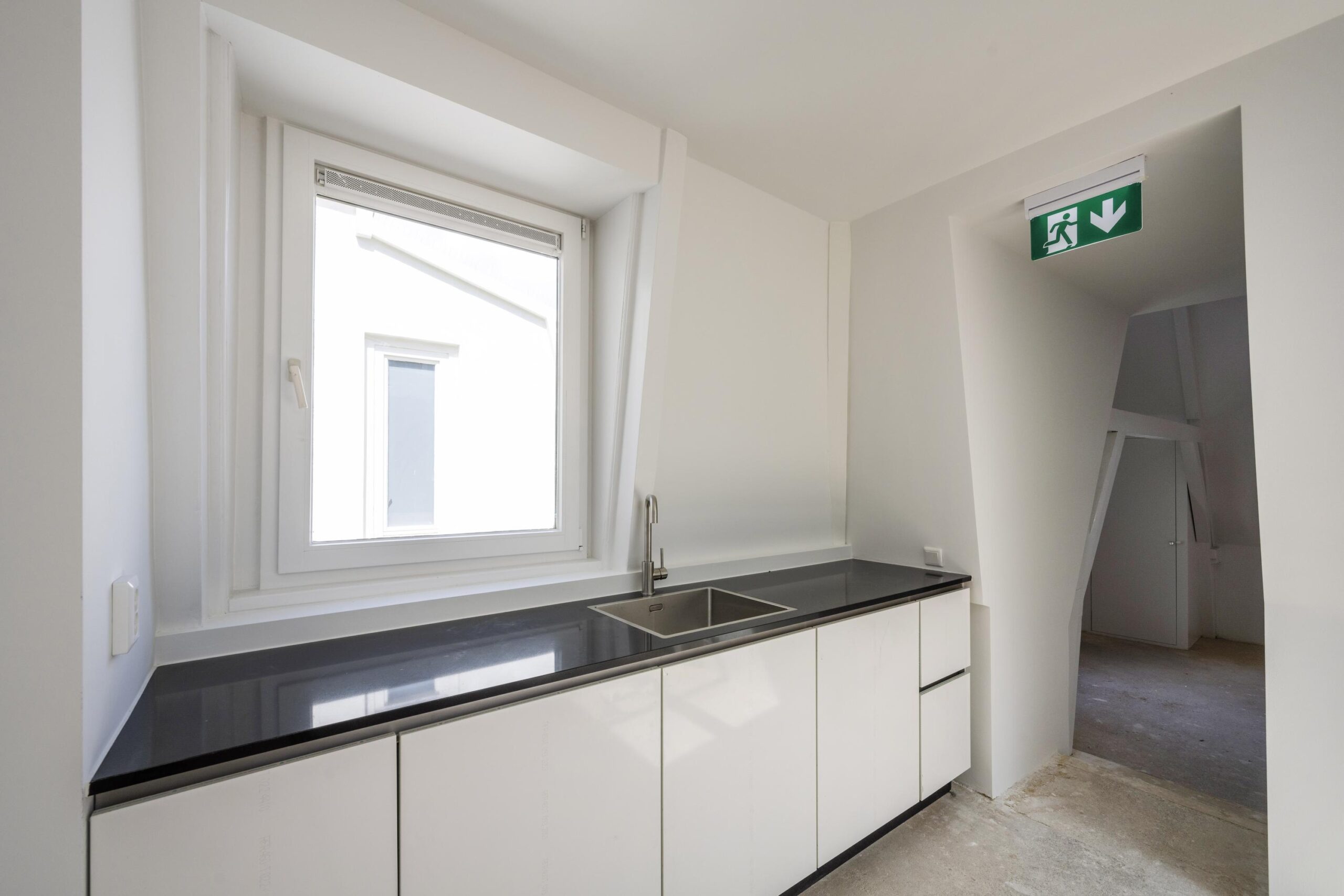 Minimalistische kitchenette met zwart werkblad, witte kasten en een raam, gelegen aan de Damrak.