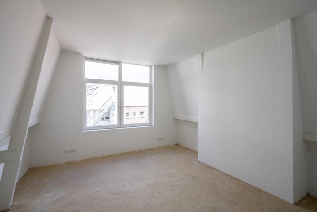 Lichte, lege zolderkamer met witgeschilderde wanden en een groot raam met uitzicht op Damrak.