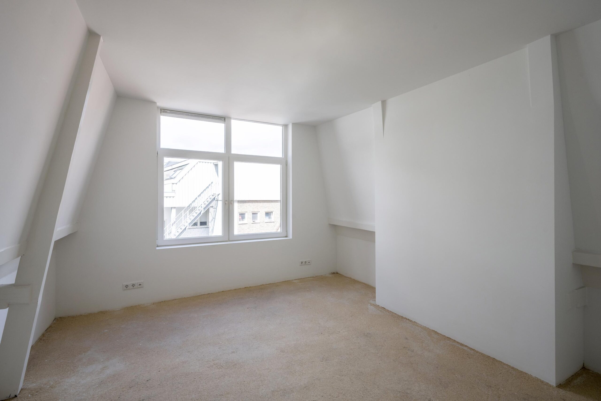 Lichte, lege zolderkamer met witgeschilderde wanden en een groot raam met uitzicht op Damrak.