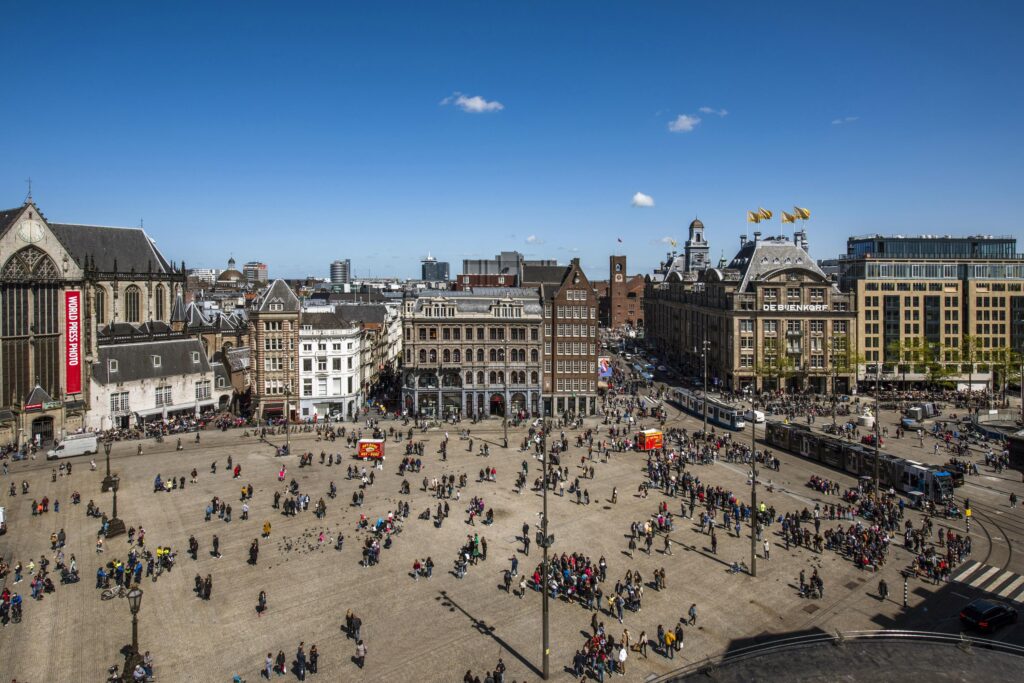 Druk Damrak in Amsterdam met uitzicht op de Dam, De Bijenkorf en de Nieuwe Kerk op een zonnige dag.