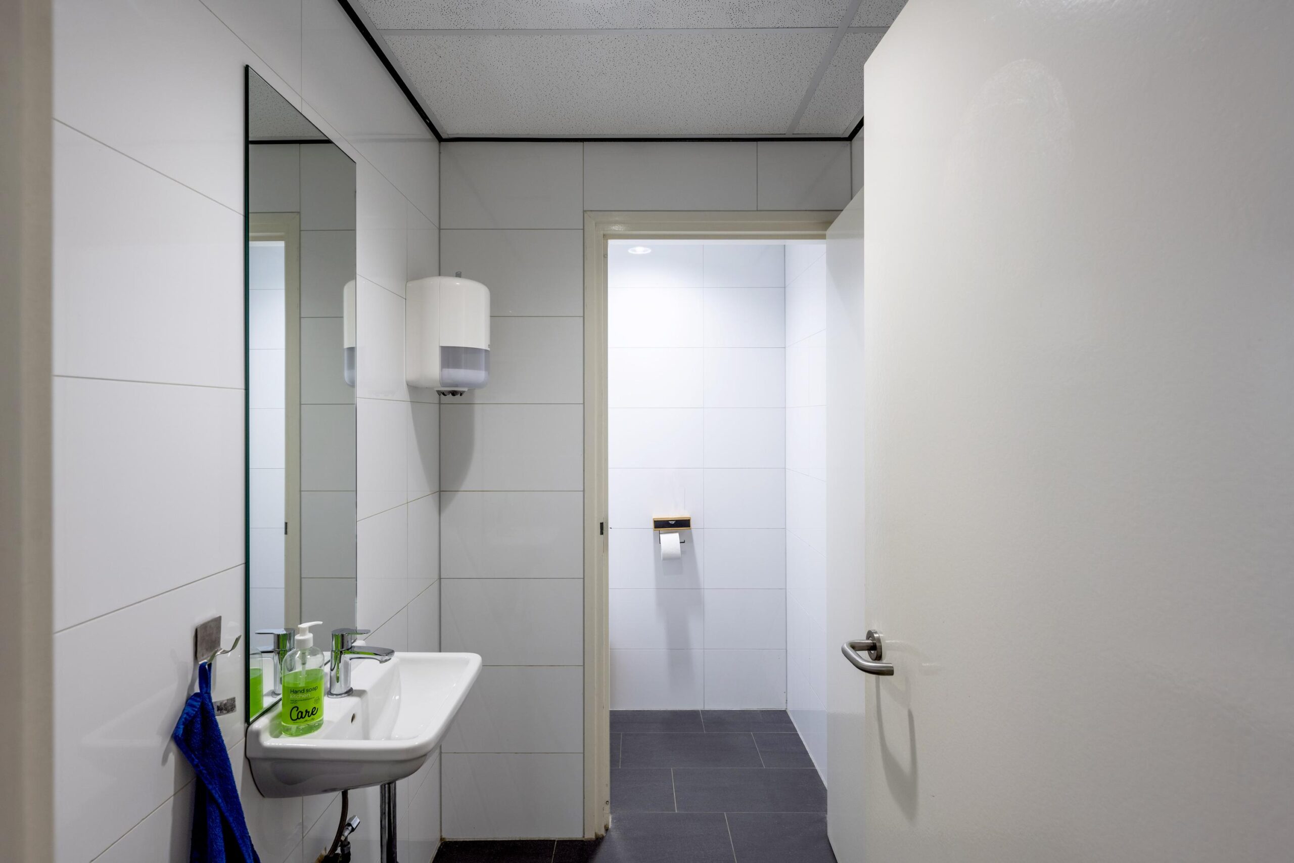 Wit betegelde toiletruimte met wasbak, spiegel, zeepdispenser en open deur naar het toilet met toiletpapierhouder.