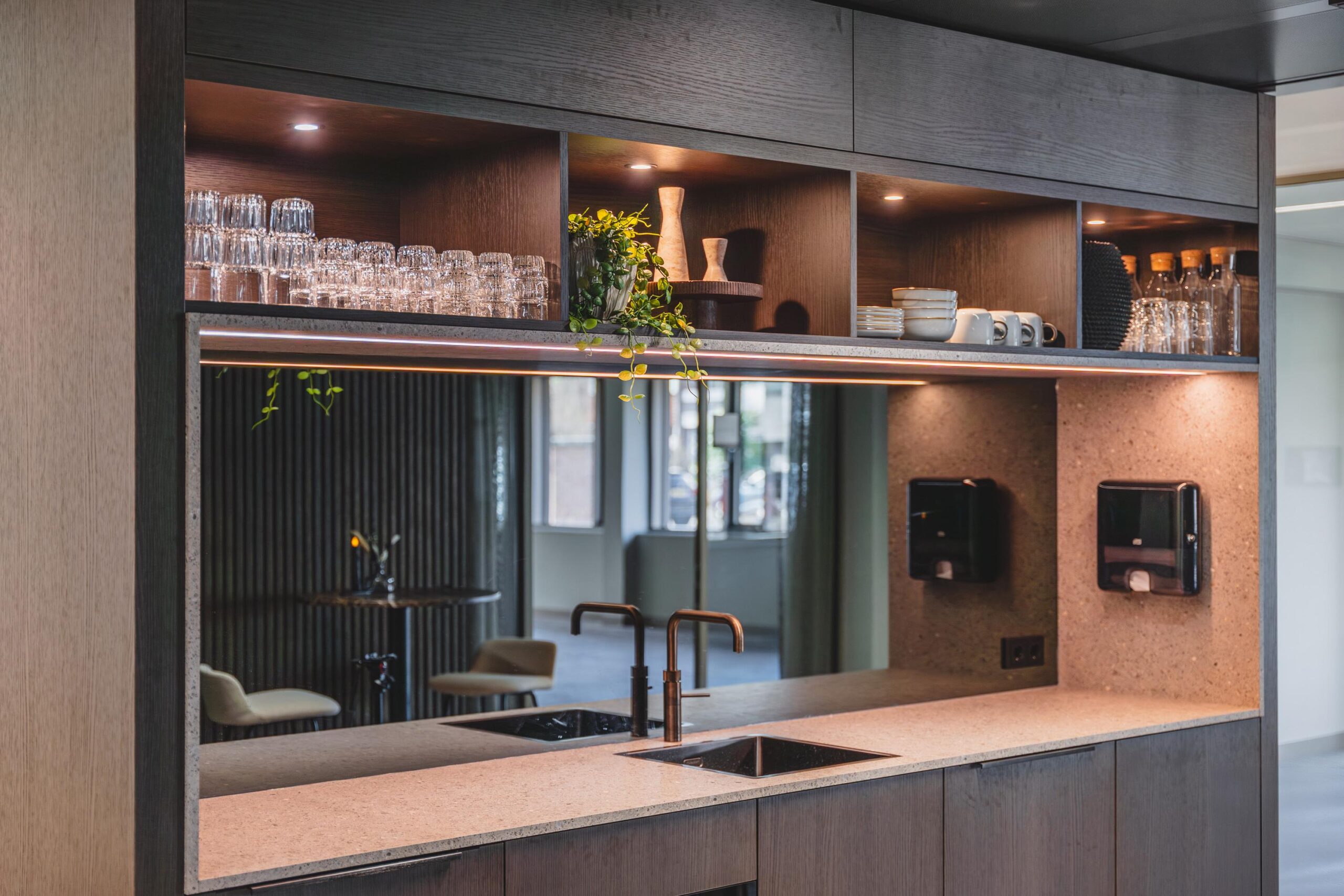Modern kitchenette with open shelves displaying glasses, cups, and decor above a counter with a sink and mirrored backsplash.