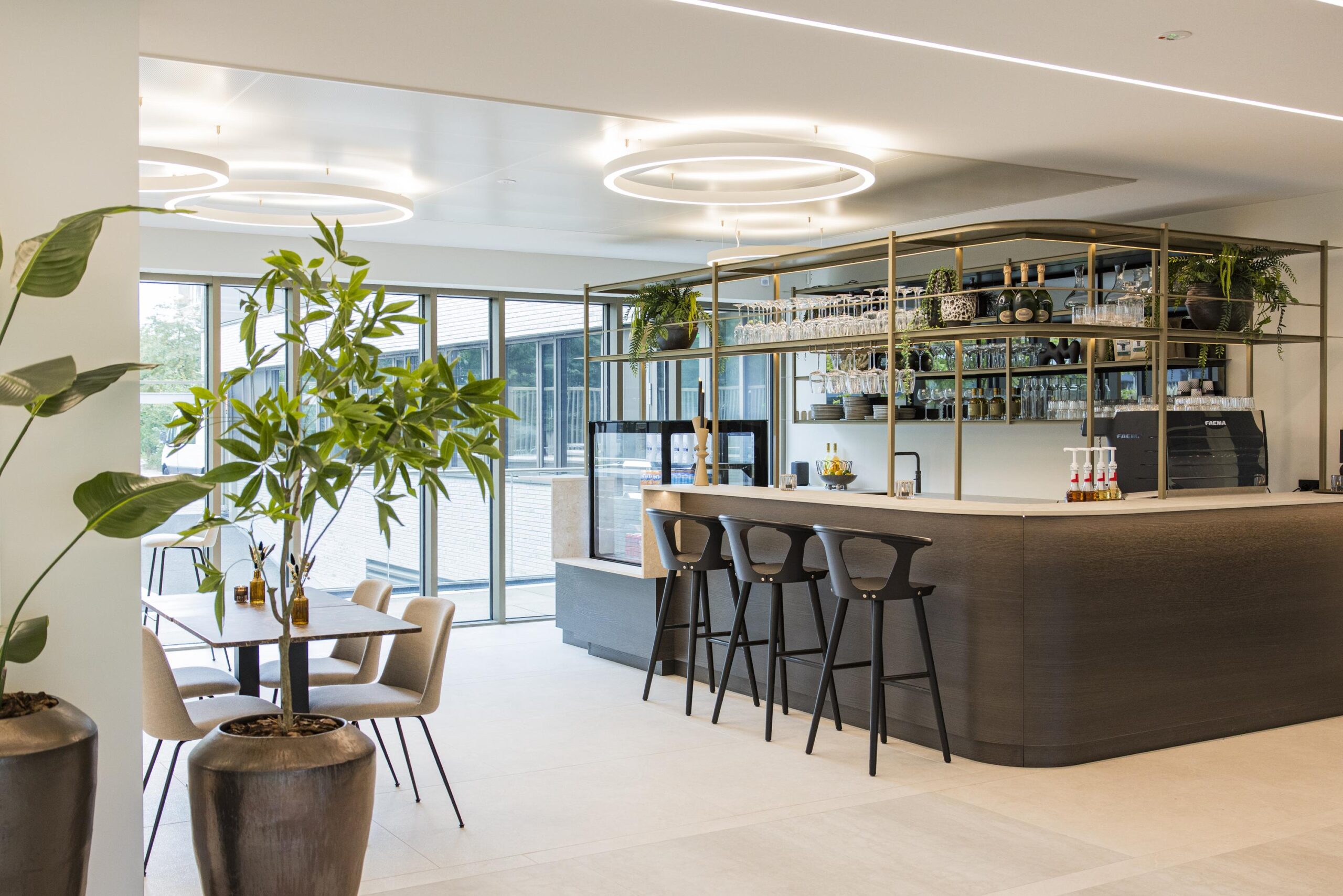 Modern hotel bar area with high stools, hanging glass racks, and indoor plants in a bright, minimalist setting.