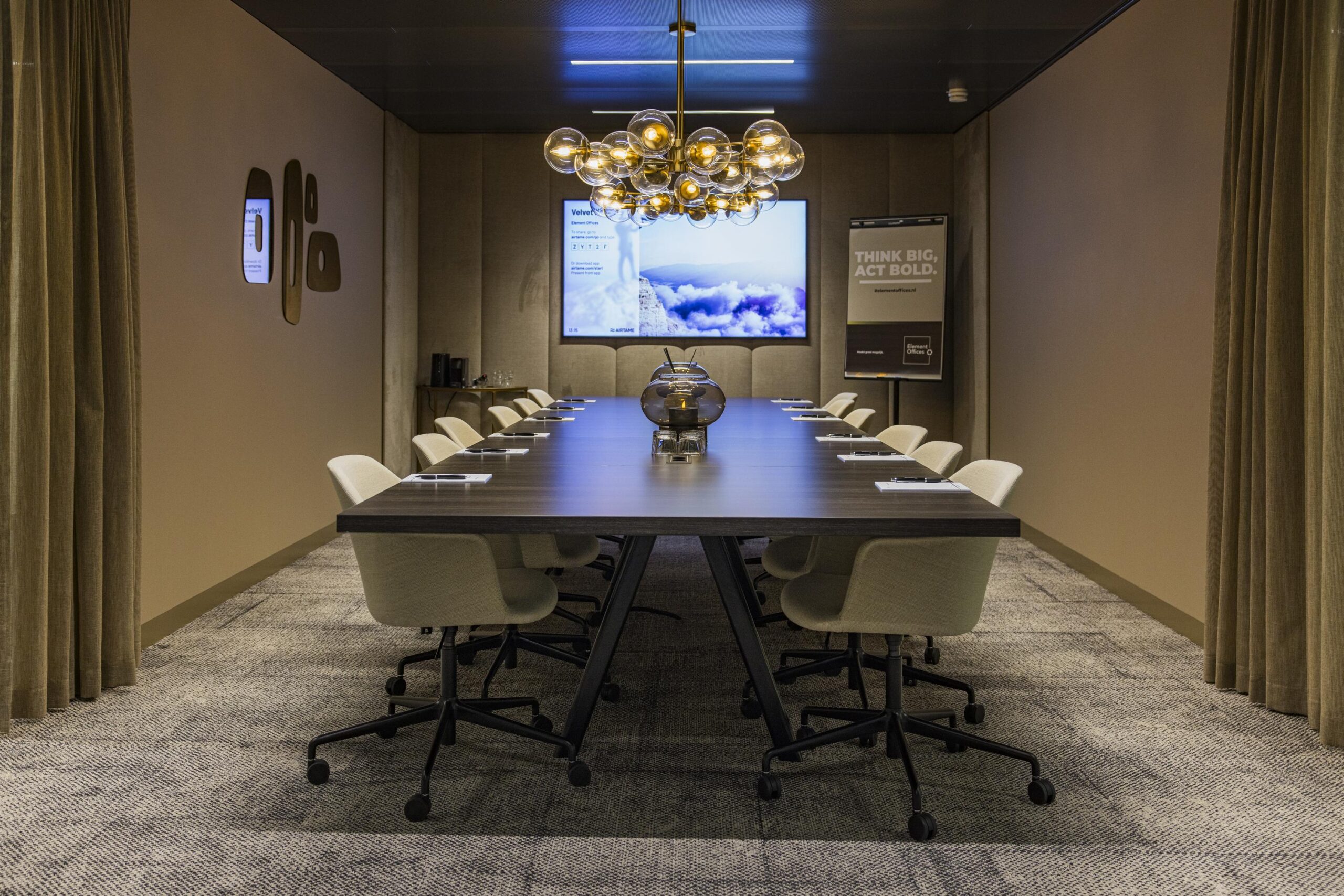 Modern conference room with a long table, beige chairs, a decorative chandelier, and presentation screens.
