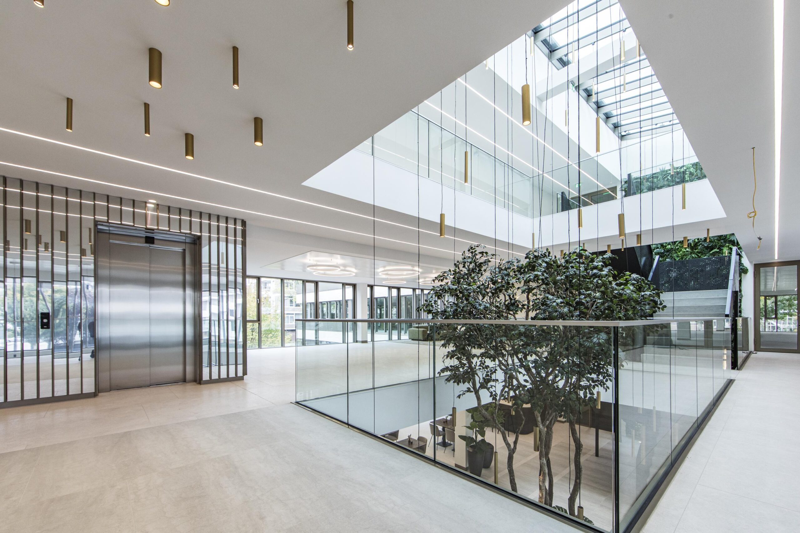Modern office atrium with glass walls, indoor trees, a skylight, and an elevator.