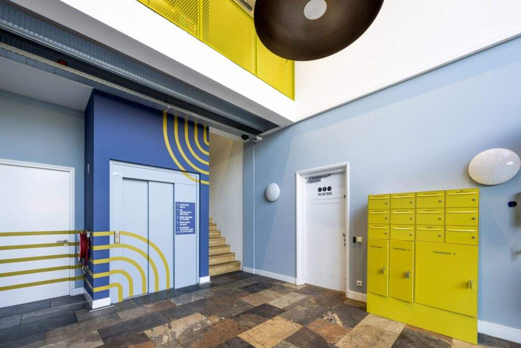 Modern entrance hall with blue and yellow accents, featuring mail lockers, an elevator, and a staircase.