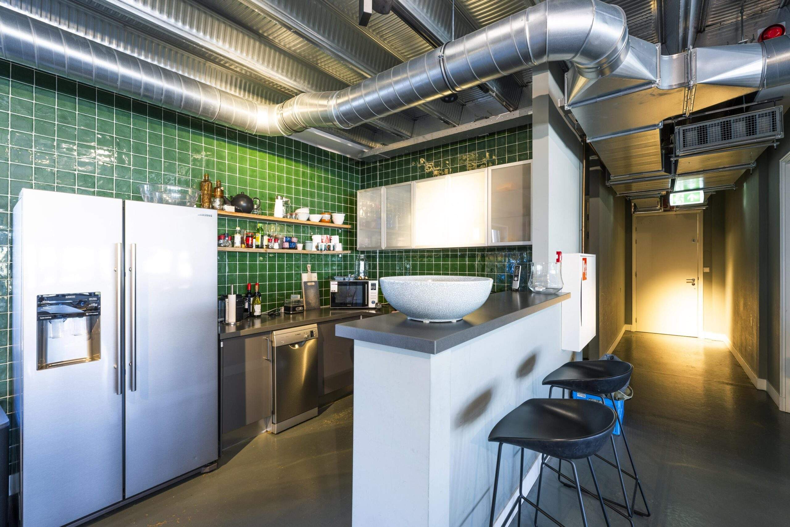 Modern industrial kitchen with green tiled walls, stainless steel appliances, and exposed ventilation ducts.