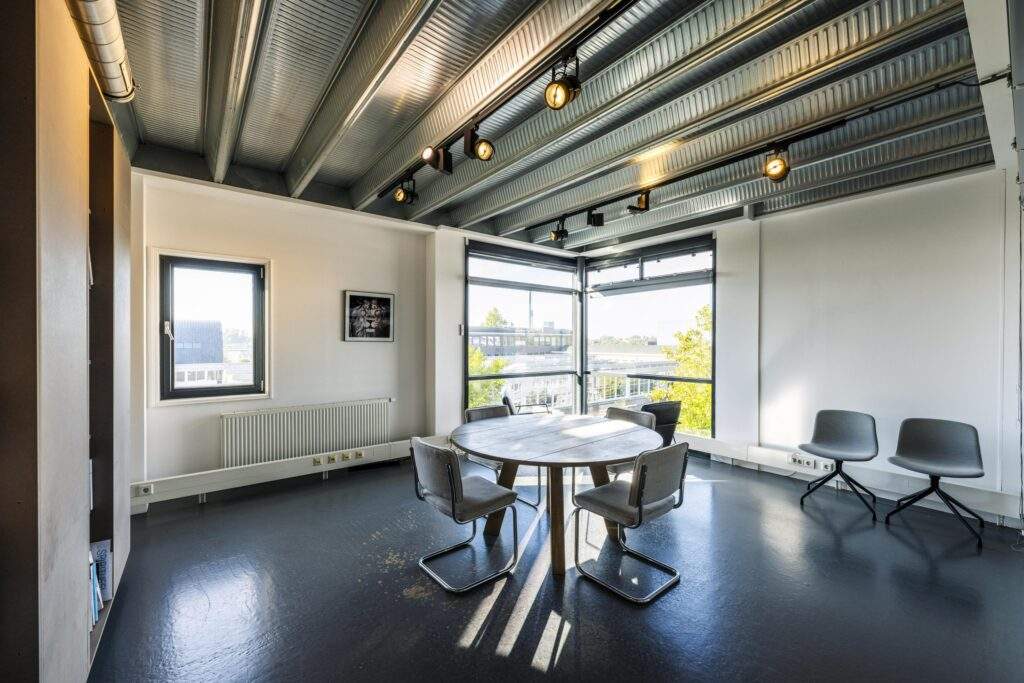 Modern meeting room on Helicopterstraat with a round table, five chairs, industrial ceiling, and large windows offering natural light.