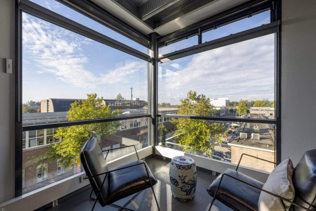 Corner seating area with two black chairs and a decorative ceramic stool, overlooking an urban street through large glass windows on Helicopterstraat.