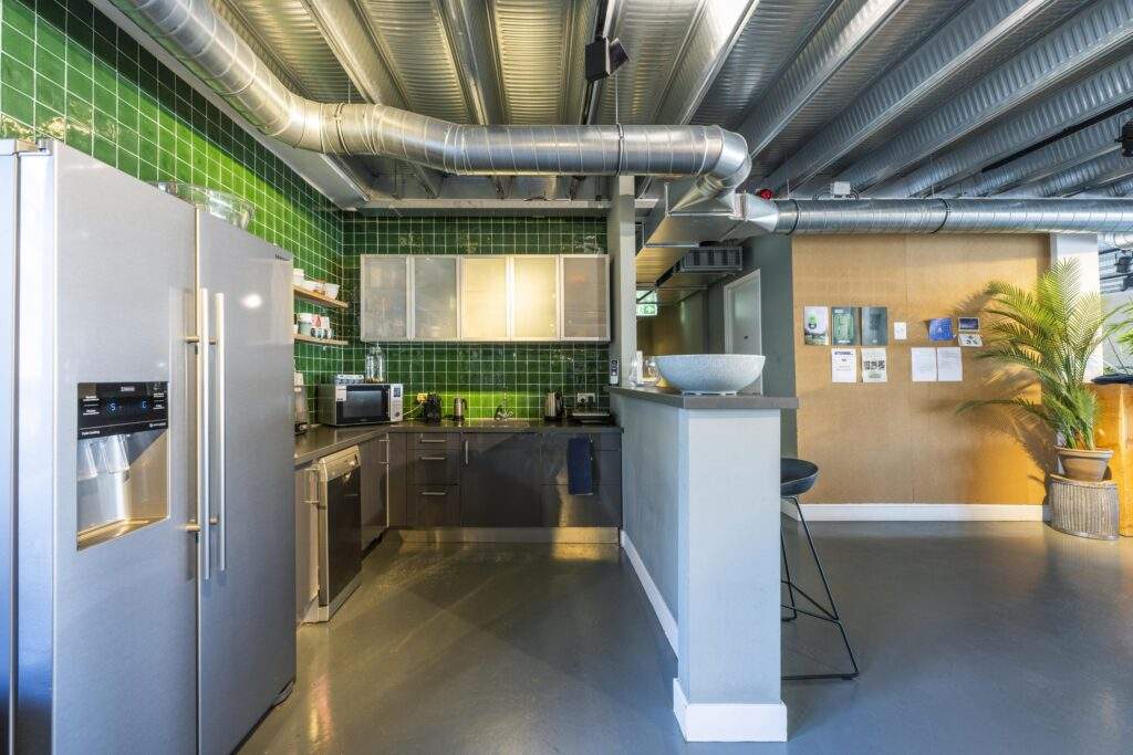 Modern industrial-style office kitchen with green tiled walls, stainless steel appliances, and exposed ventilation ducts.