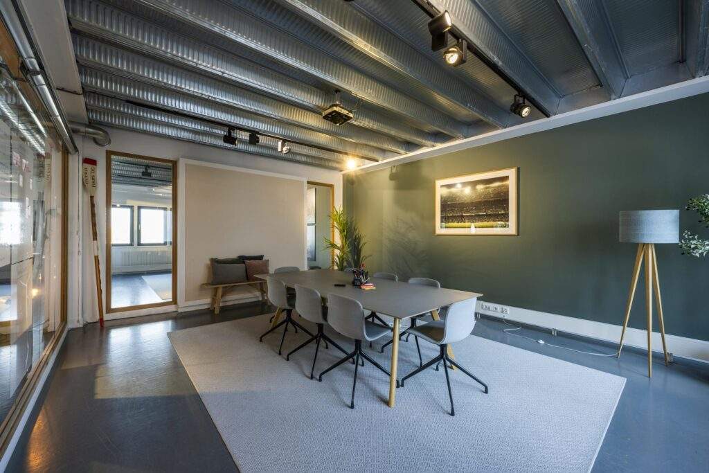 Modern meeting room with a long table, eight chairs, a green accent wall, and industrial ceiling lighting.