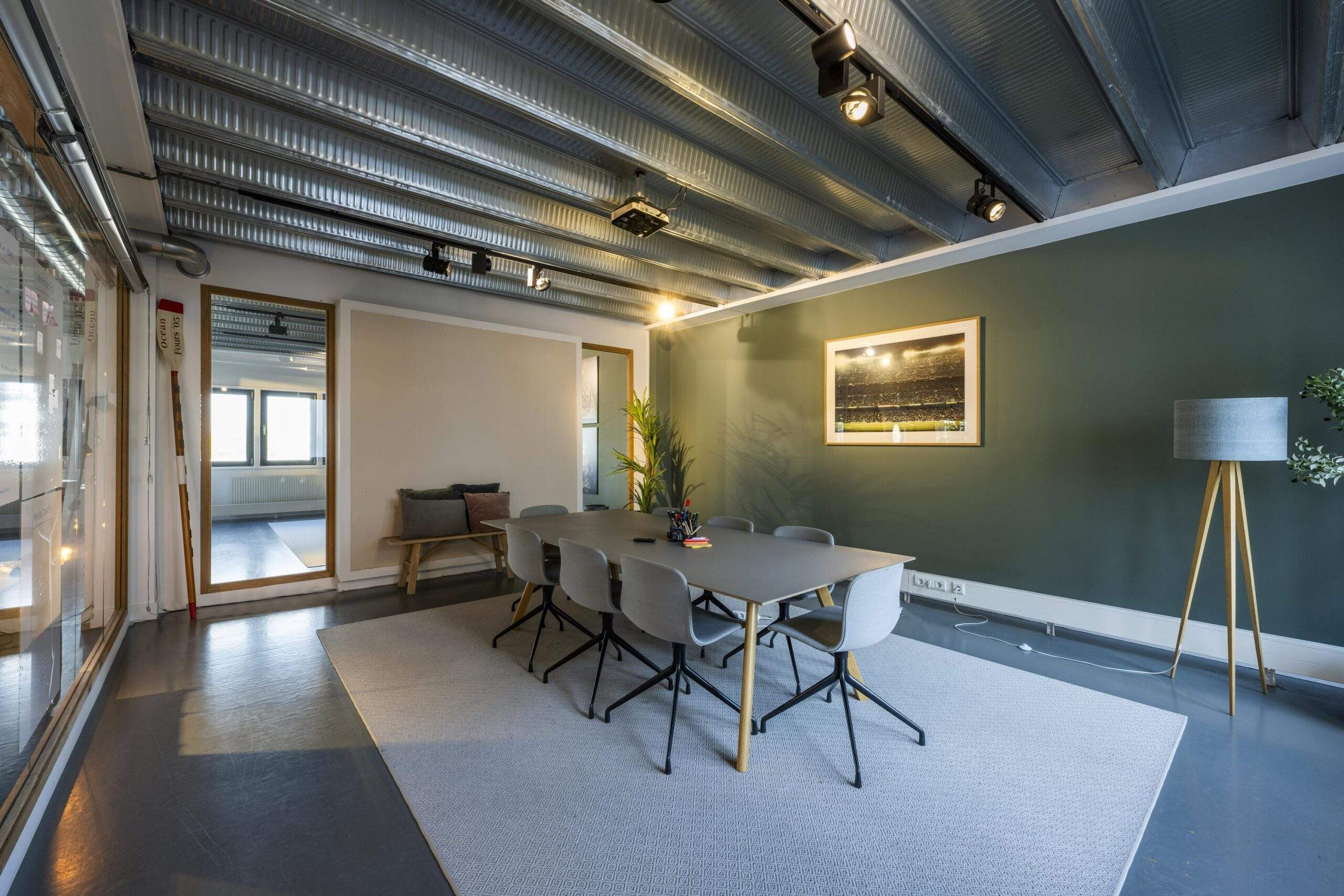 Modern meeting room with a long table, eight chairs, a green accent wall, and industrial ceiling lighting.