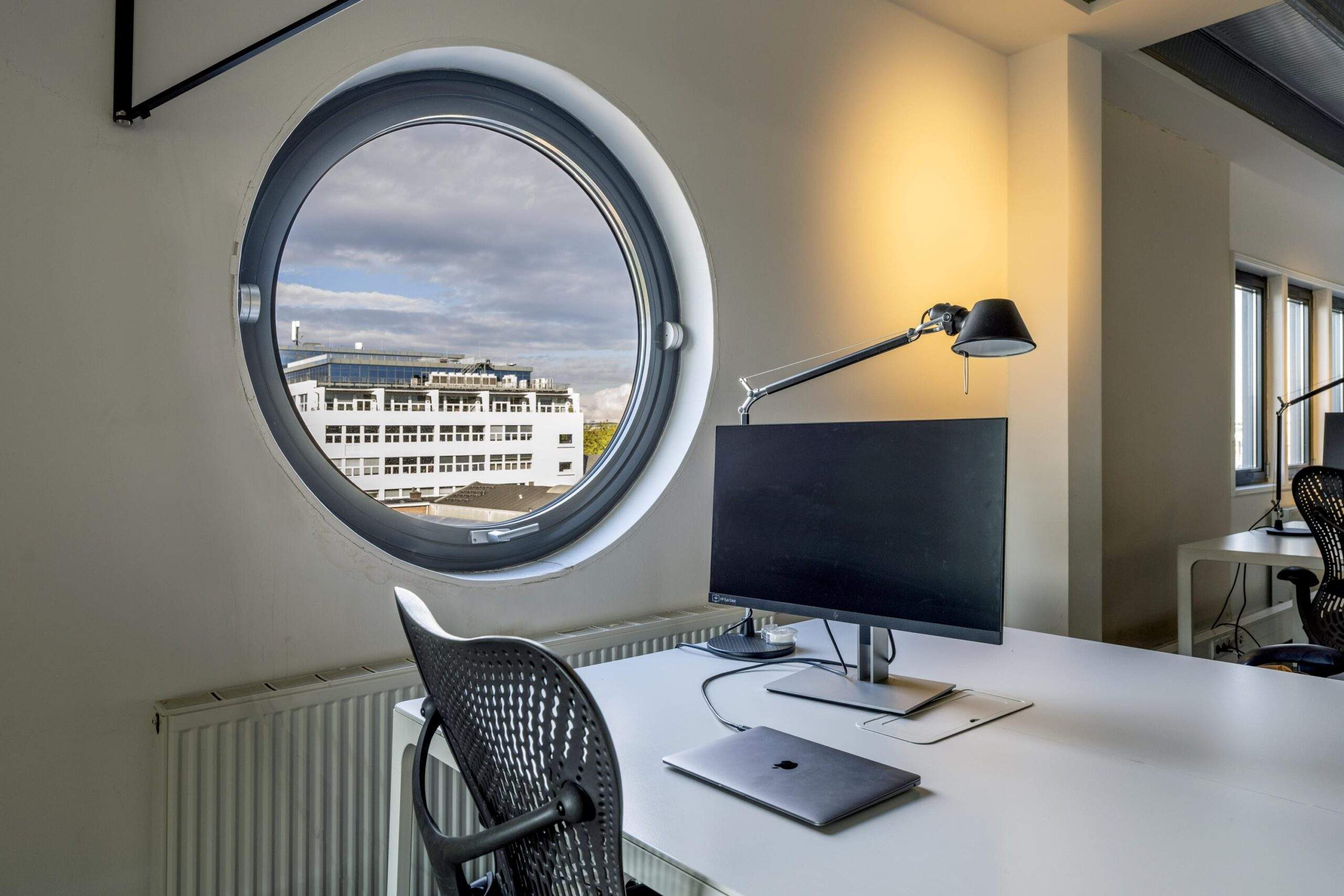 Modern office workspace with a round window overlooking a white building on Helicopterstraat.