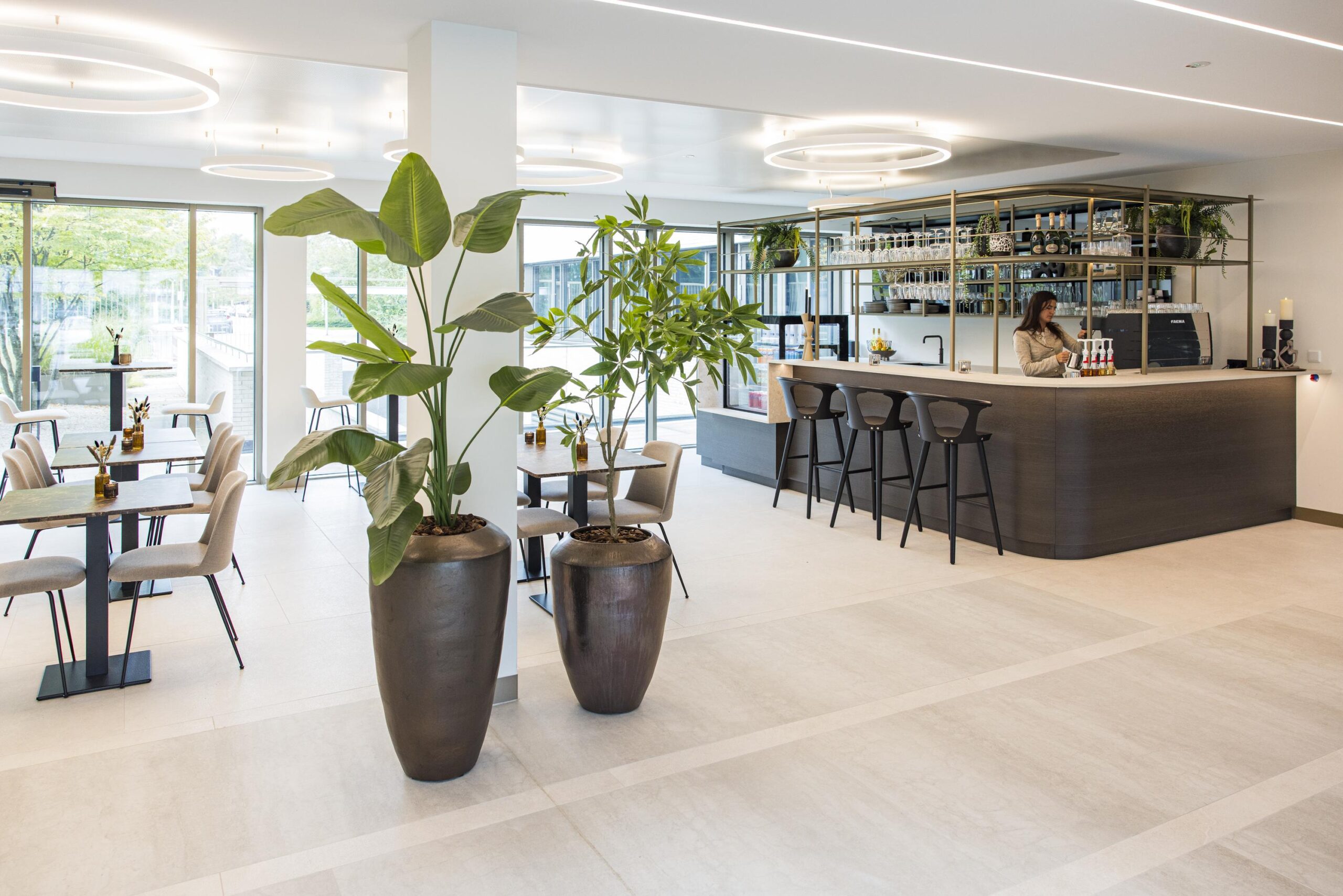 Modern interieur van een lichte horecaruimte in Walborg met een bar, zitplaatsen, grote planten en een vrouwelijke medewerker achter de bar.