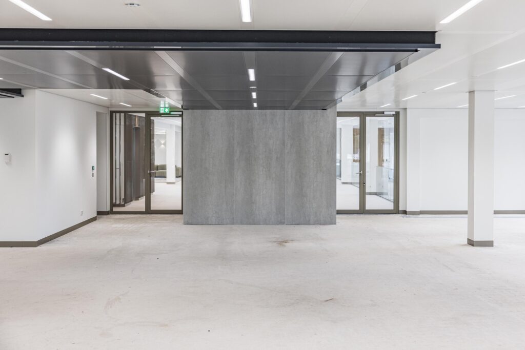 Leeg kantoorpand met glazen deuren, witte muren en een grijze scheidingswand in het midden.