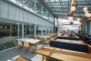 Modern en licht restaurantinterieur aan de Amstelveenseweg met houten tafels, glazen wanden en sfeerverlichting.
