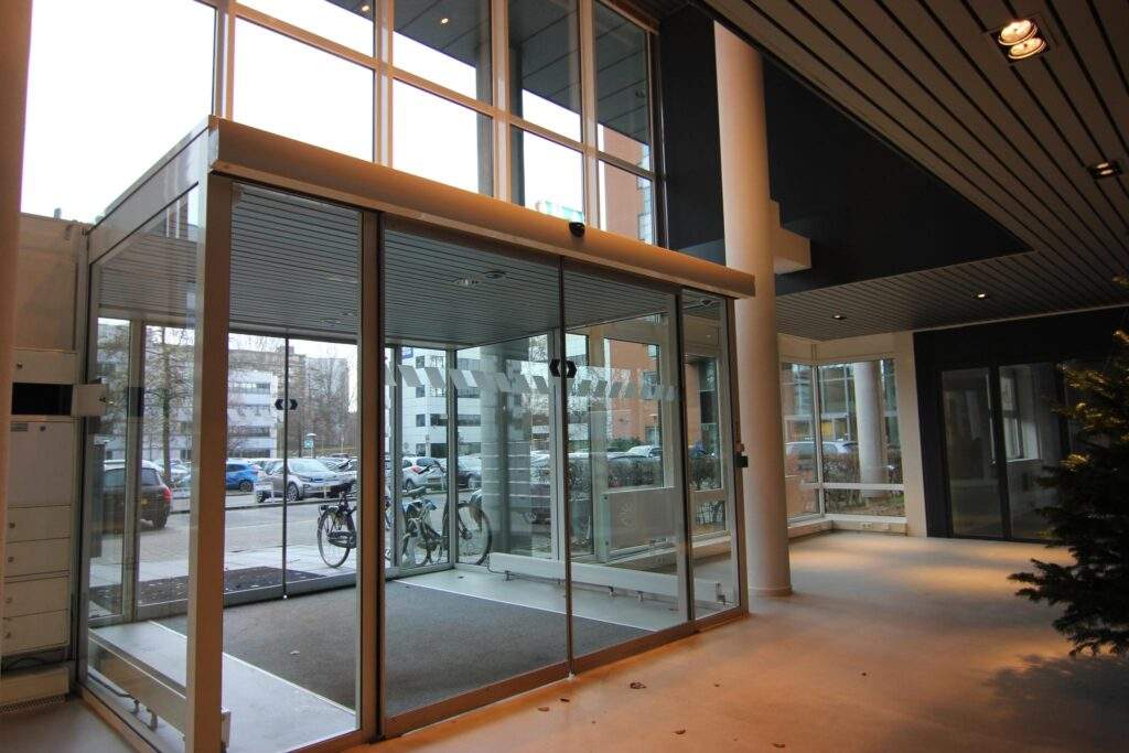 Glass entrance with automatic sliding doors and a view of parked bicycles and cars outside.
