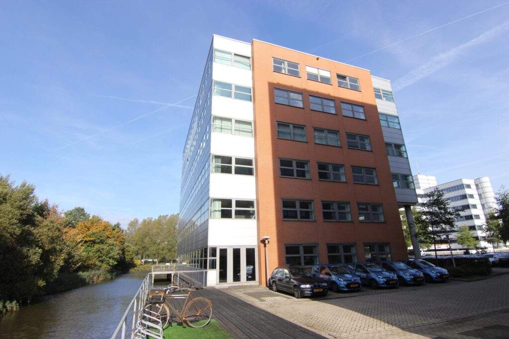 Modern office building named Entrada located next to a canal, with parked cars and bicycles in front.