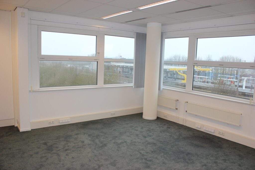 Empty office space with large windows, grey carpet, and a view of a nearby train station.
