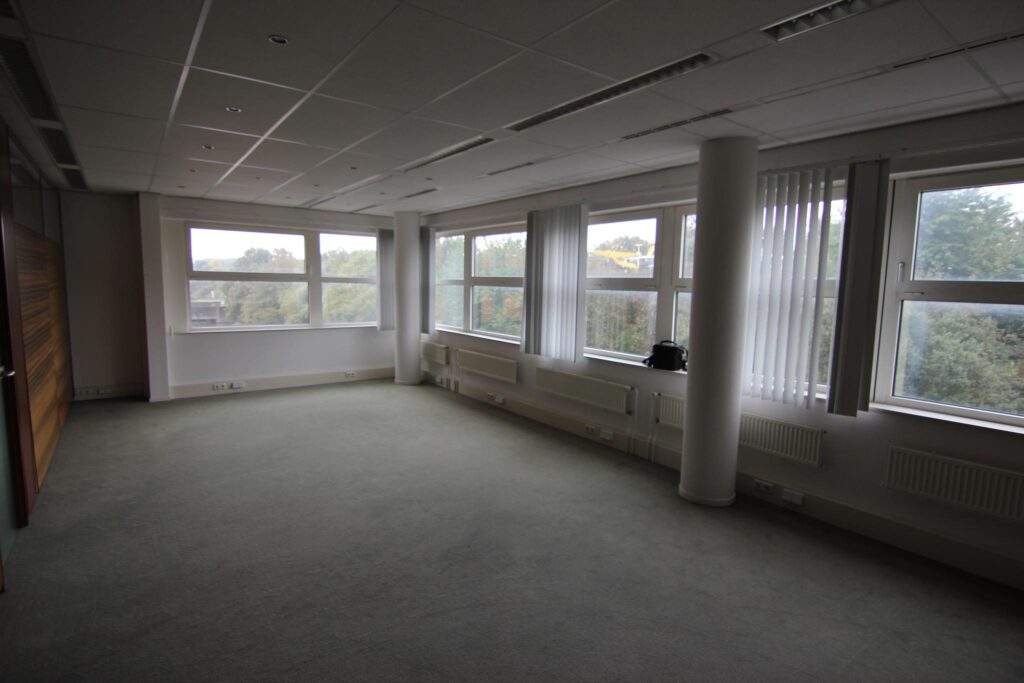 Empty office space with large windows, white walls, and grey carpeting.
