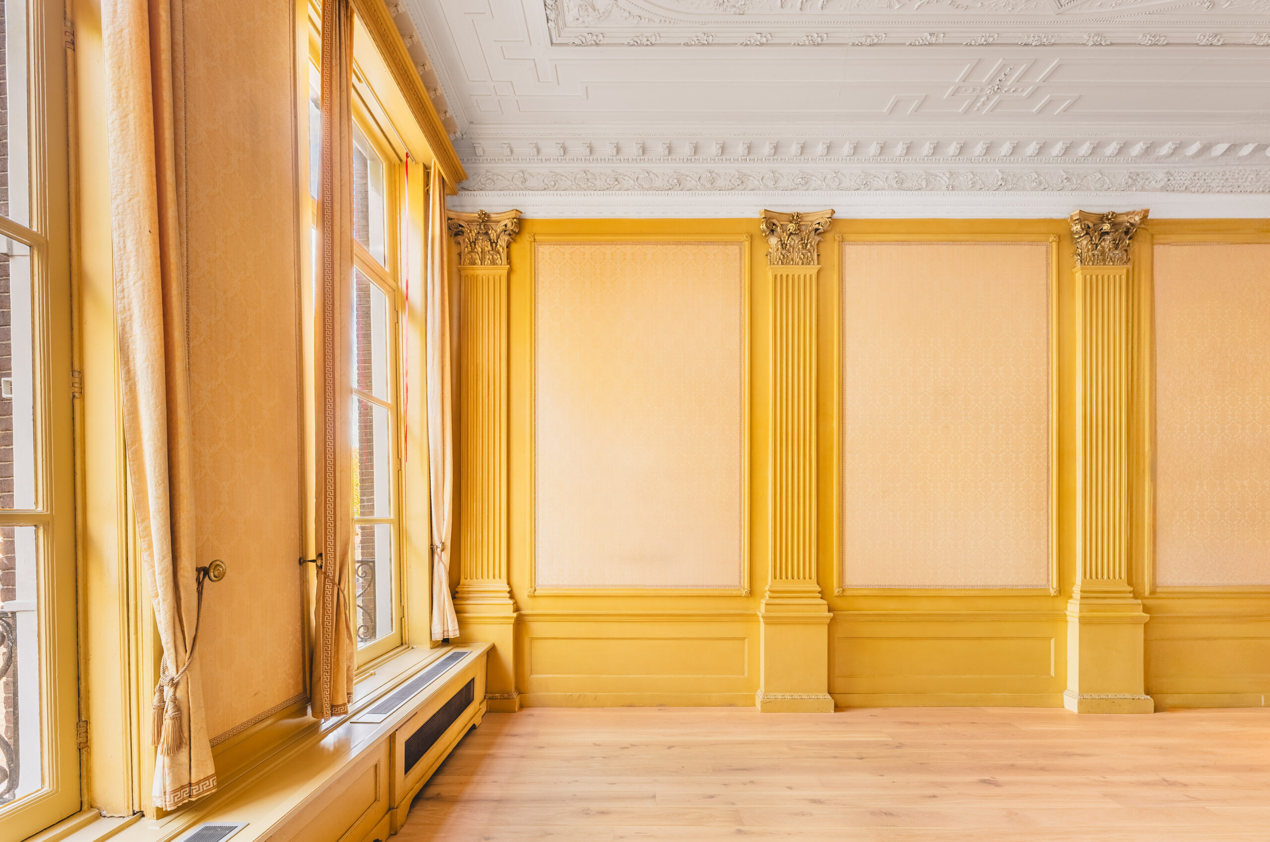 Elegant yellow room with large windows, classical wall paneling, and an ornate white ceiling.