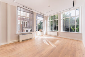 Bright empty room at Adress_12 with large windows, light wooden floor, and views of greenery and a brick building.