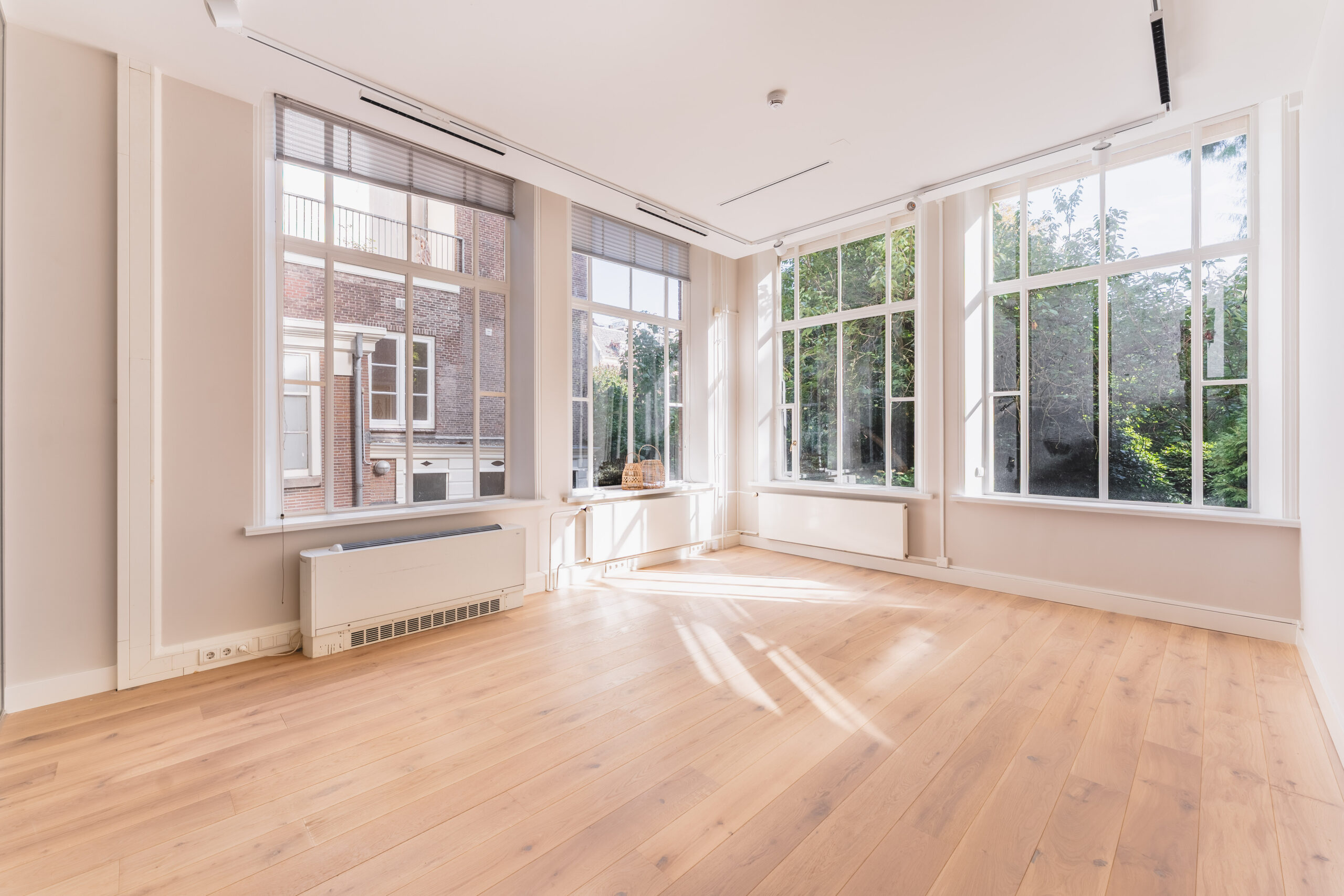 Bright empty room at Adress_12 with large windows, light wooden floor, and views of greenery and a brick building.