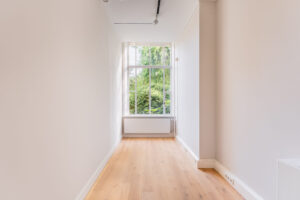 Bright, narrow room with light wooden flooring and a large window overlooking green trees.