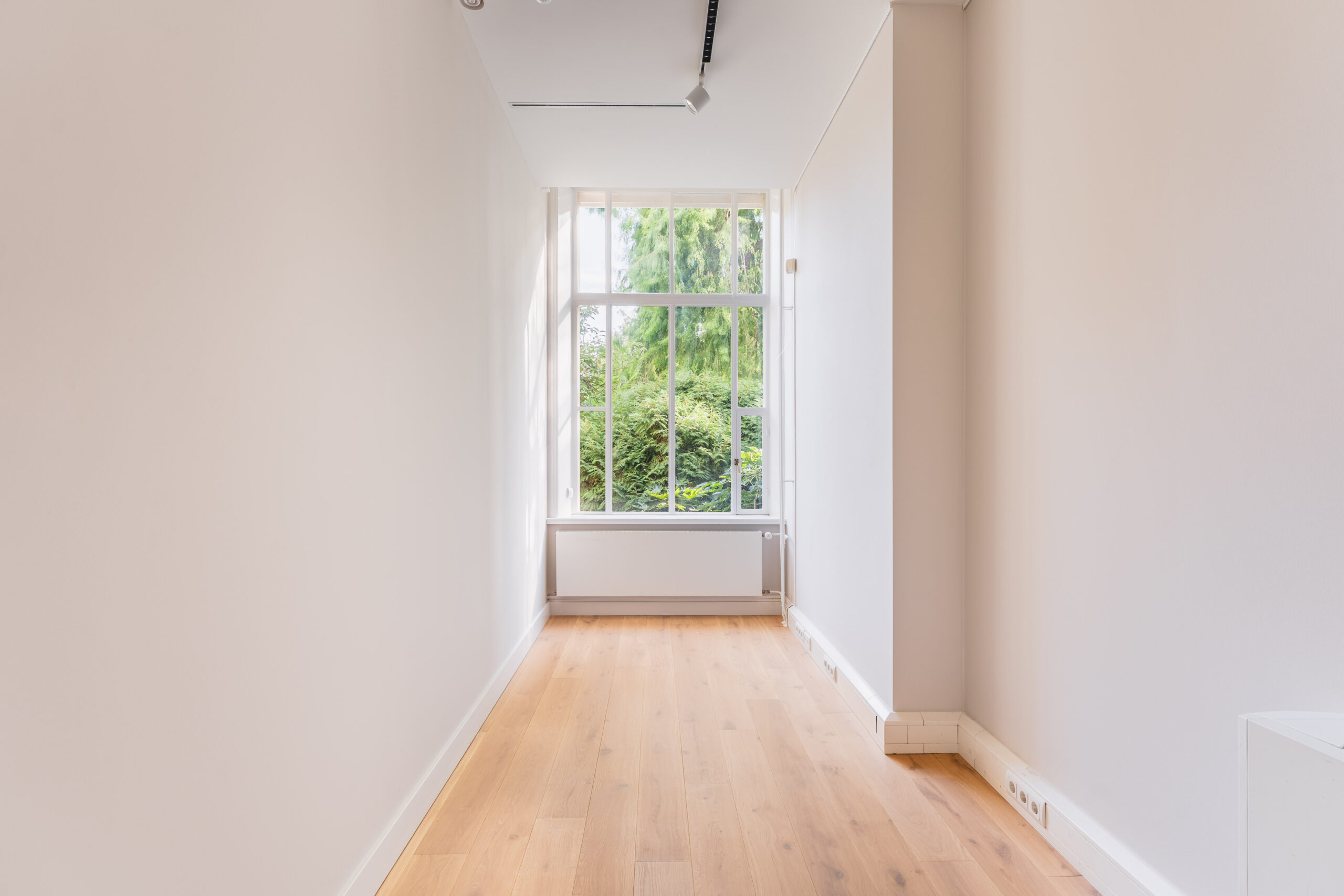 Bright, narrow room with light wooden flooring and a large window overlooking green trees.
