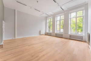 Bright, empty room at Adress_15 with large windows, light wooden floor, and white walls.