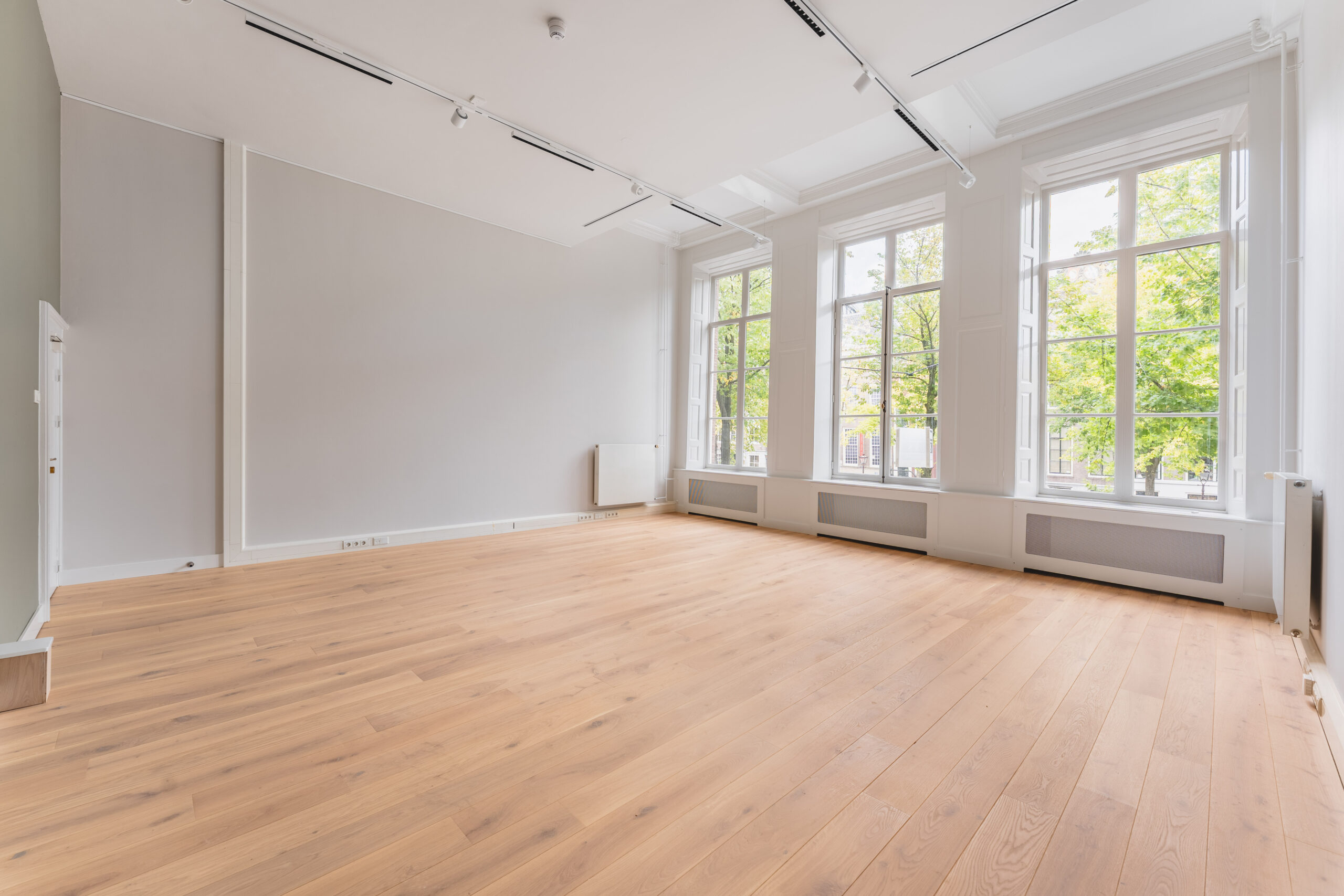 Bright, empty room at Adress_15 with large windows, light wooden floor, and white walls.
